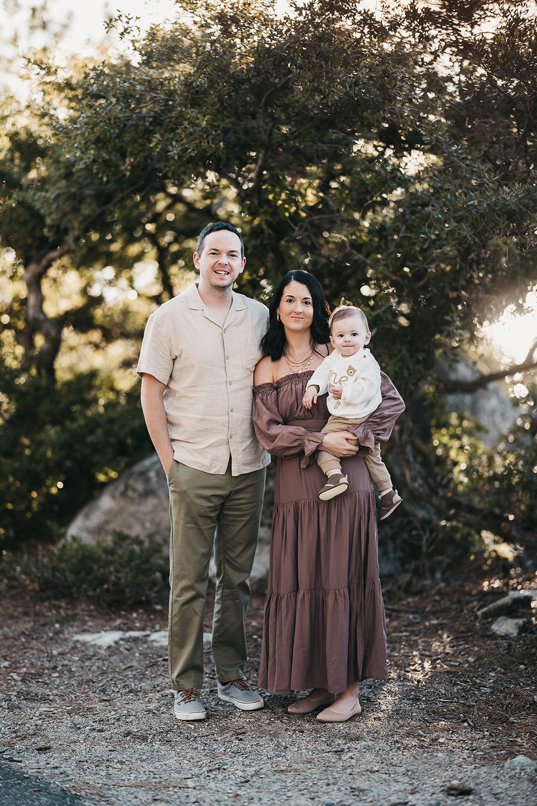 A family of three standing outdoors during sunset, with trees in the background. The man is wearing a beige shirt and khaki pants, the woman is in a long brown dress holding a young child dressed in cream and brown, all smiling at the camera.