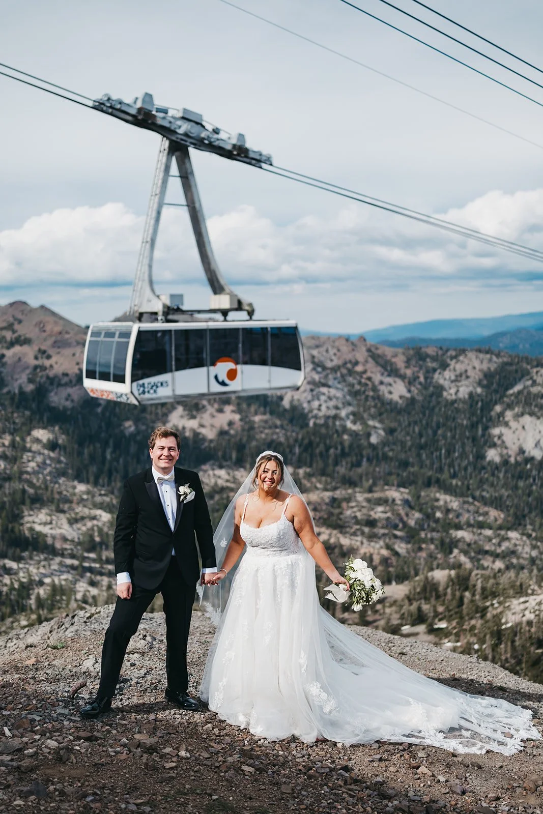 A bride and groom in wedding attire standing outdoors on a rocky hill with mountains in the background, holding hands and smiling, with a cable car above them in the sky.
