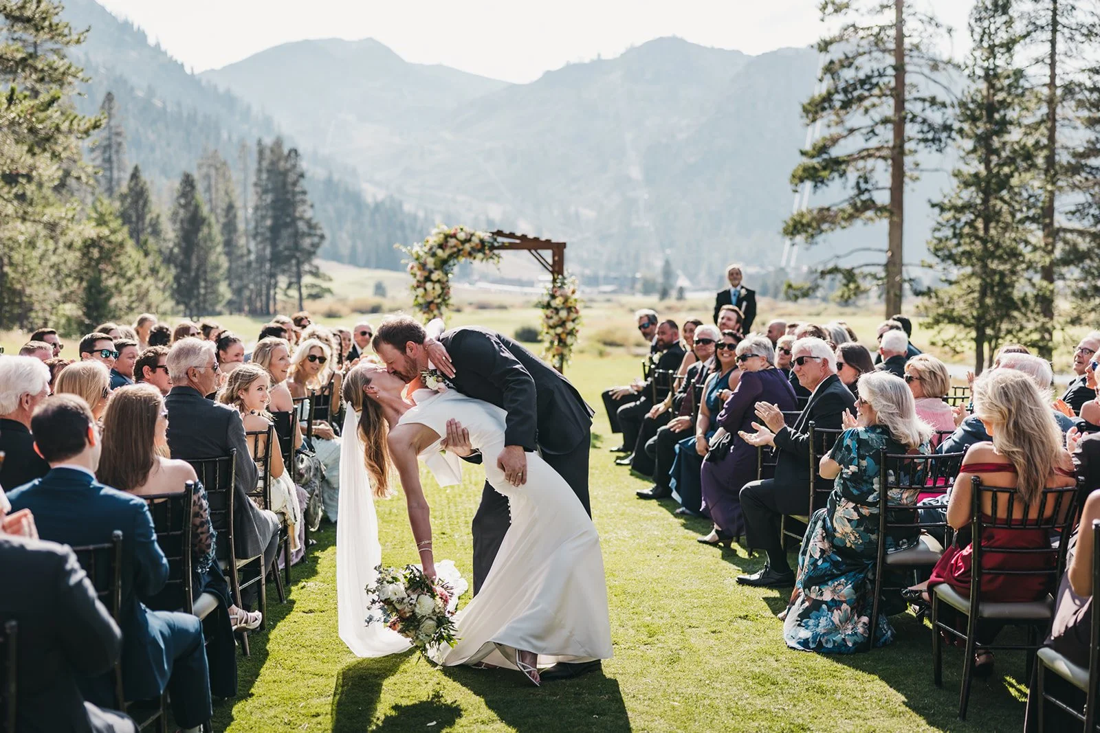 A bride and groom kiss during an outdoor wedding ceremony in a mountain setting at Everline Resort, surrounded by seated guests on a grassy field with trees and mountains in the background.