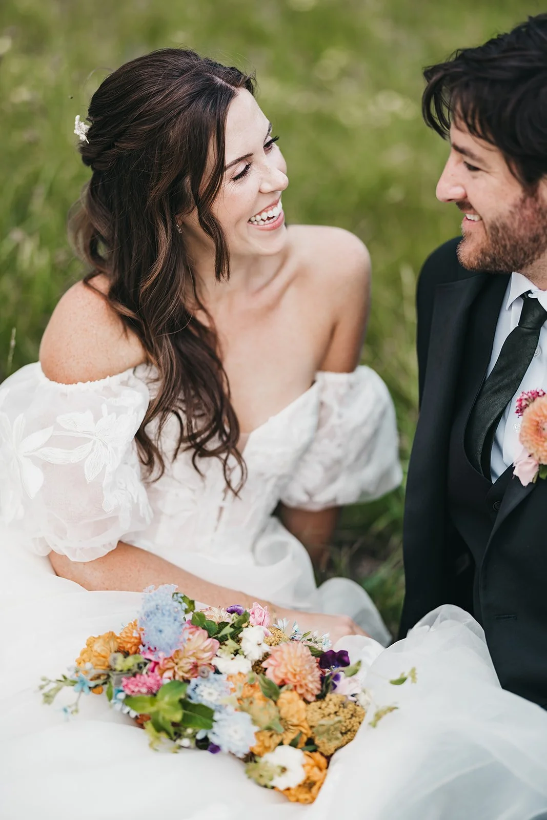 A bride and groom sitting on grass  at Kirkwood Mountain Resort with the bride smiling and holding a colorful bouquet, dressed in wedding attire, outdoors.