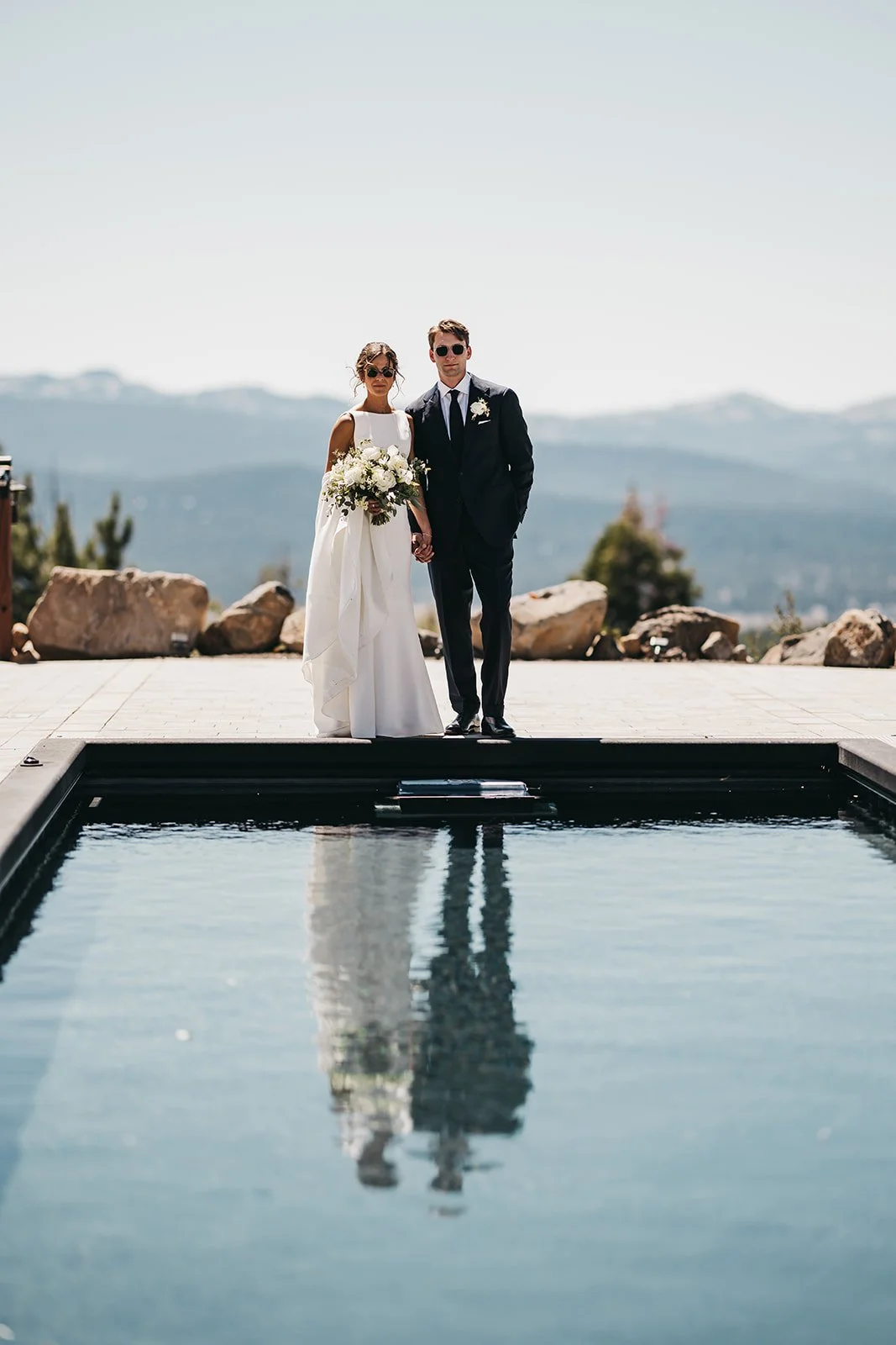 A bride and groom standing on a platform by a reflecting pool, holding hands, with mountains in the background, during a wedding ceremony outdoors on a sunny day.