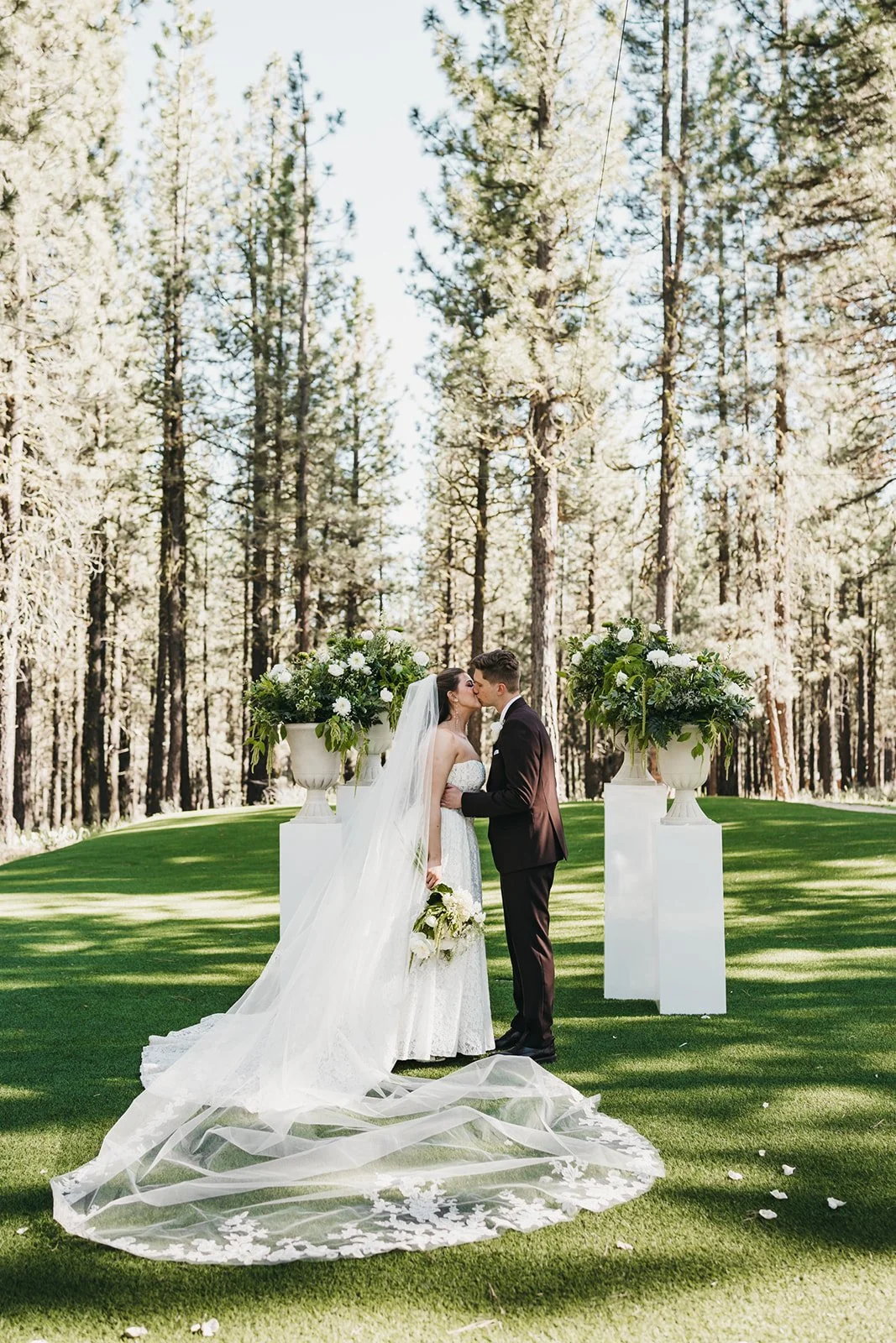A bride and groom sharing a kiss during their outdoor wedding ceremony in a forested area, with large floral arrangements in white urns on tall pedestals.