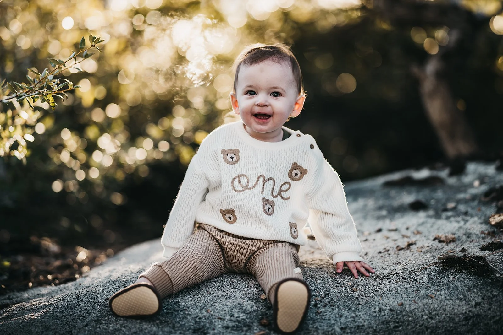 A joyful baby sitting on a rock outdoors with a blurred background of greenery and sunlight, wearing a cream sweater with bear designs and the word 'one' on it, and beige pants.