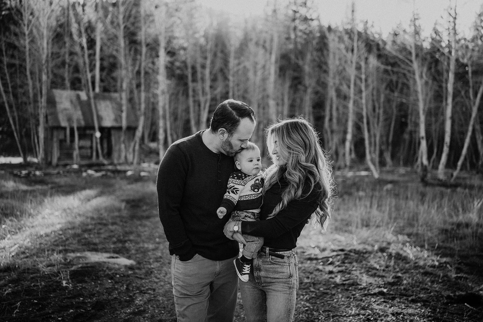 A black-and-white photo of a family of three outdoors, with trees in the background. The father and mother are gently touching foreheads with their baby, who is being held by the mother. The family is smiling and appears happy.