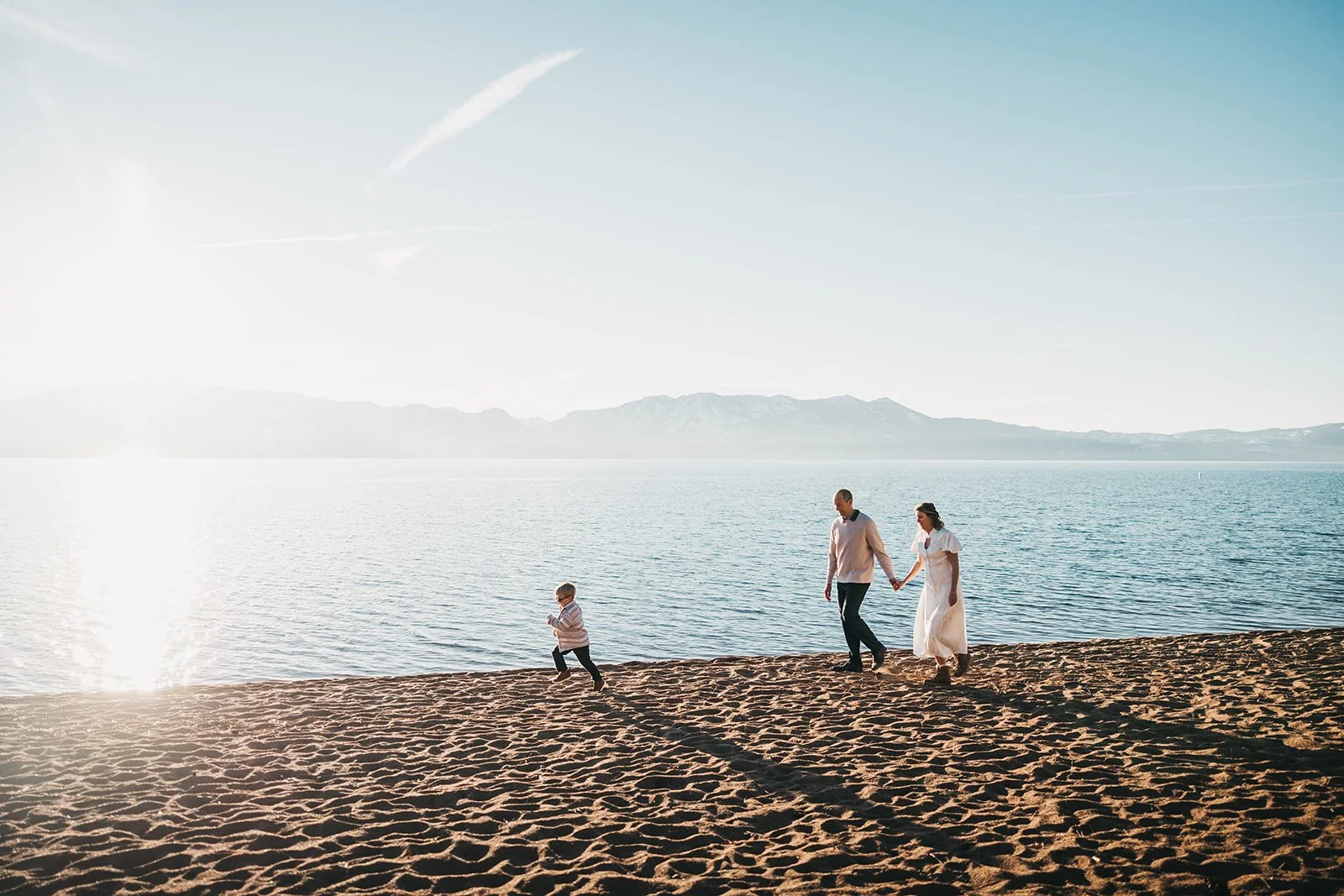 A family of three, including a young boy, walks along a sandy beach by the water during sunset, with mountains in the background.