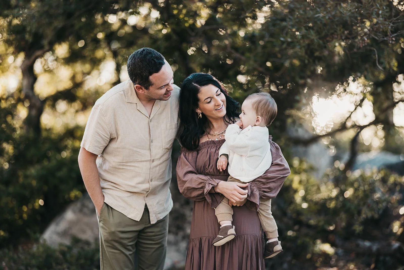A family outdoors in the evening with sunlight filtering through trees. A woman holds a young child, and a man stands beside them smiling.