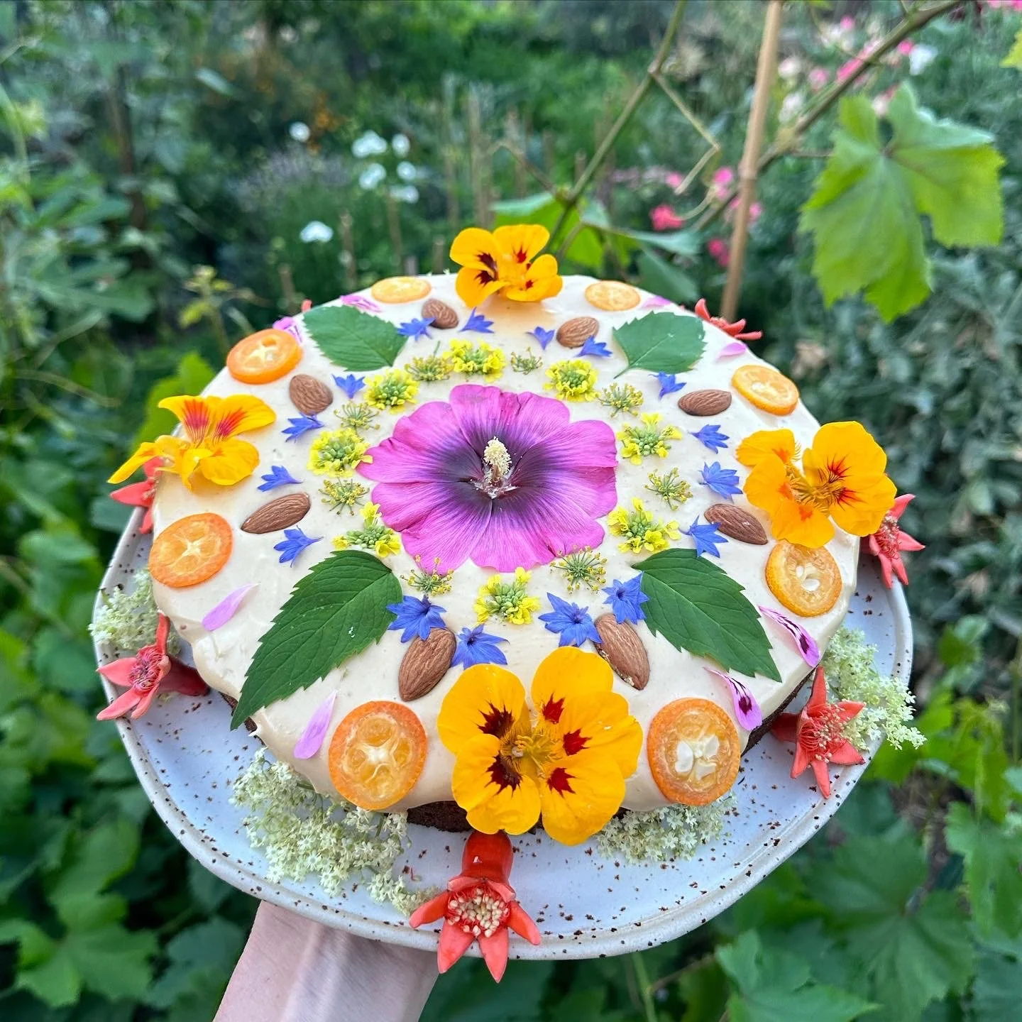 A decorated cake with colorful edible flowers, leaves, and nuts on top and around the sides, set outdoors with green foliage in the background.