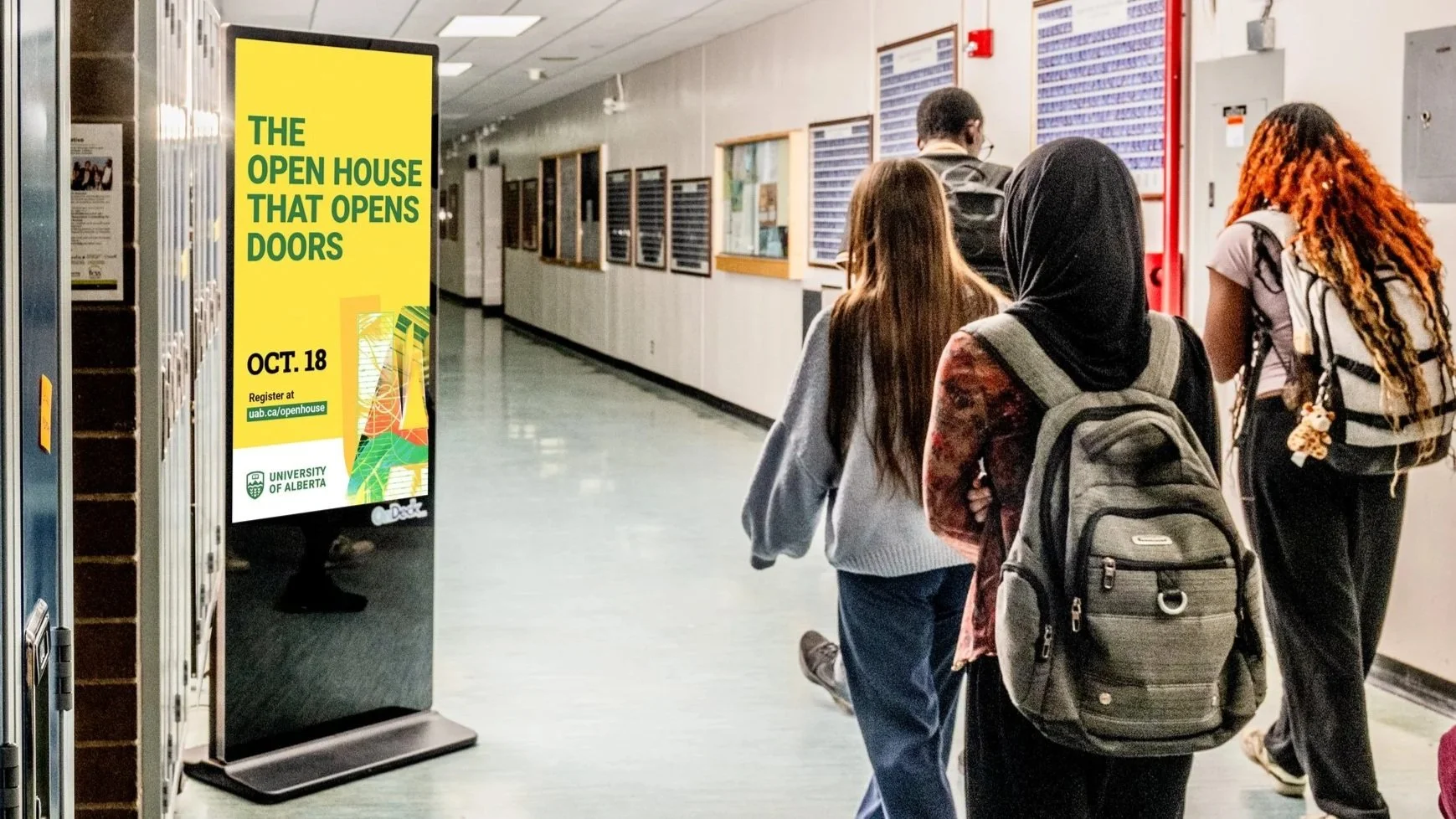 Students walking down a hallway at the University of Alberta, with a bright yellow poster on the left advertising an open house event on October 18th.