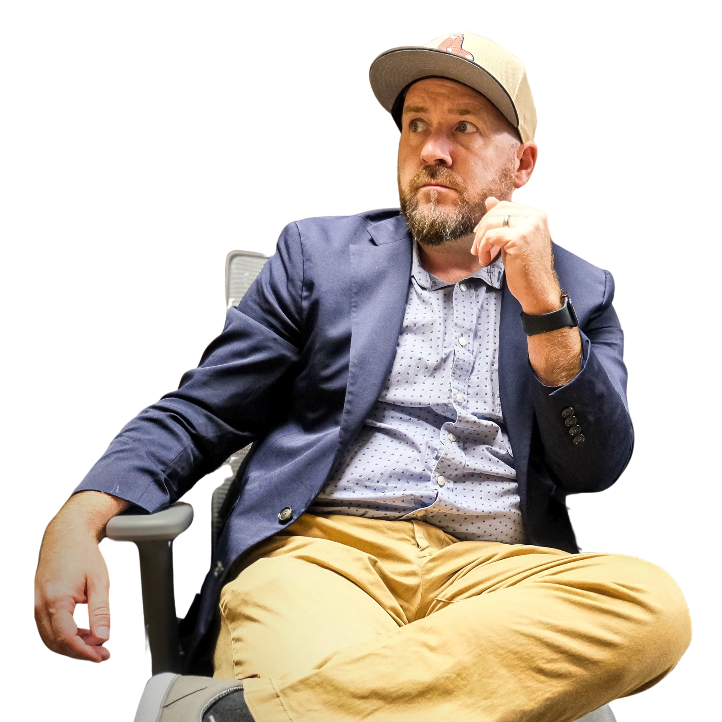 A man with light skin, a beard, and wearing a beige cap with a pink design, navy blazer, light blue patterned shirt, tan pants, and grey shoes, sitting in a chair with a white background.