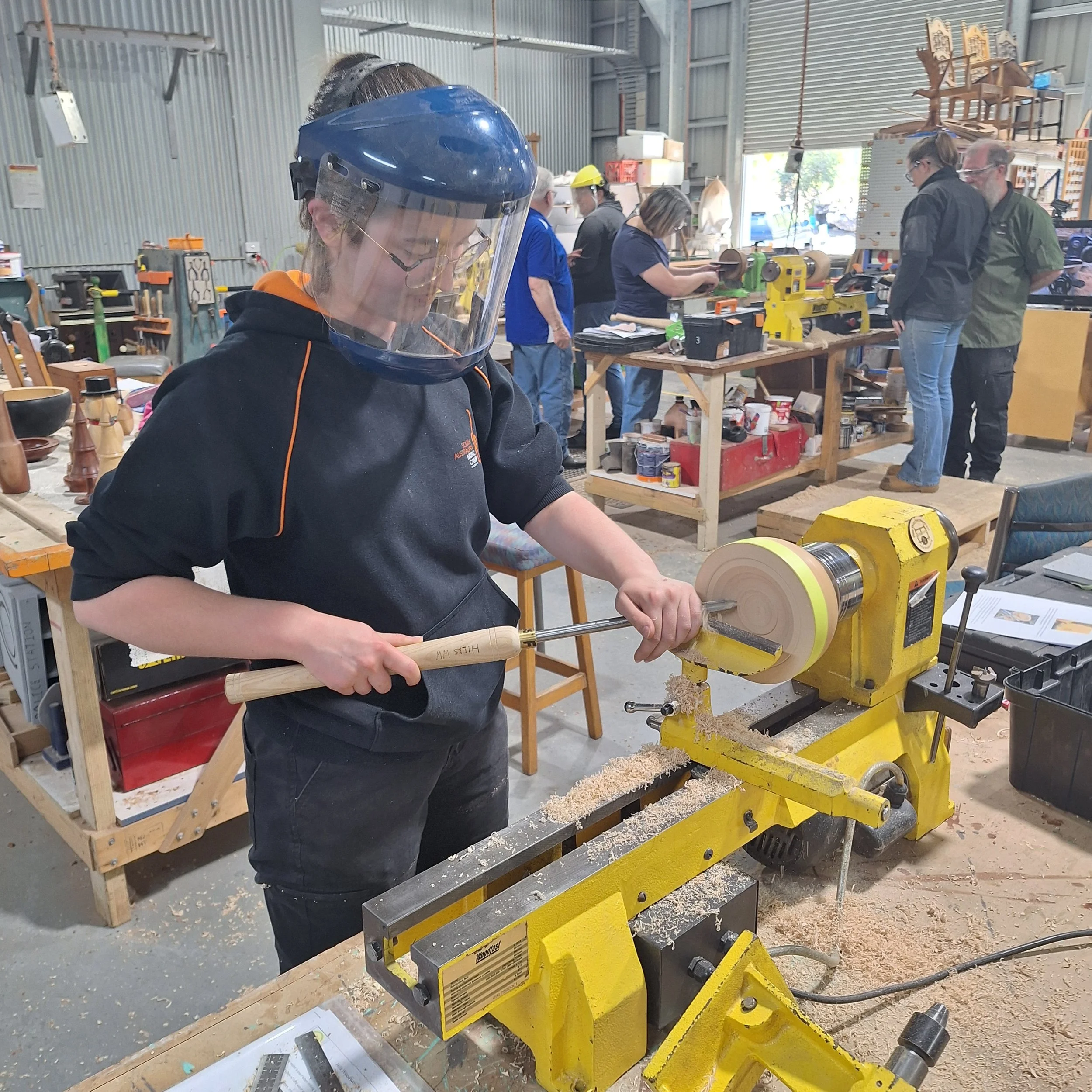 Amber stands next to a woodturning lathe. She's wearing a face shield and holding a chisel, turning a bowl.