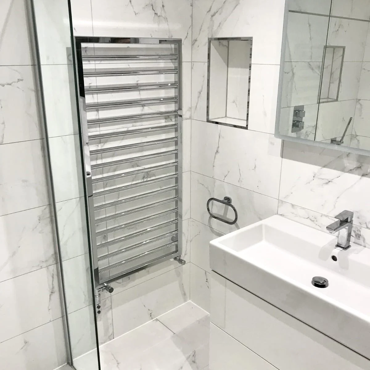 Modern bathroom with white marble walls and floor, a rectangular white sink with a chrome faucet, a large mirror above the sink, a chrome towel ring, and a chrome heated towel rack.
