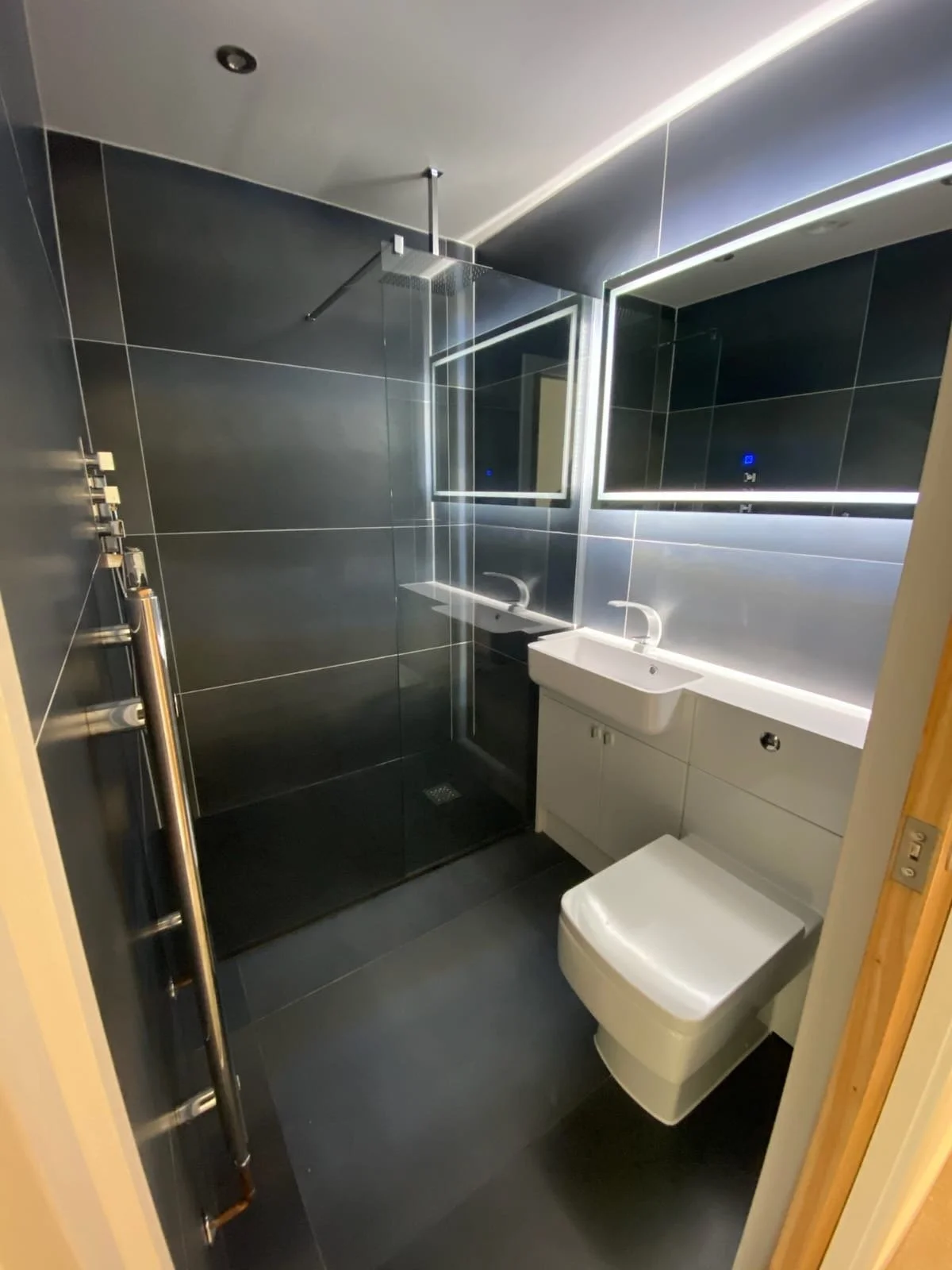 Modern bathroom with dark gray tiled walls and floor, a glass shower enclosure with a rainfall showerhead, a white vanity with integrated sink, a large mirror with LED lighting, and a wall-mounted toilet.