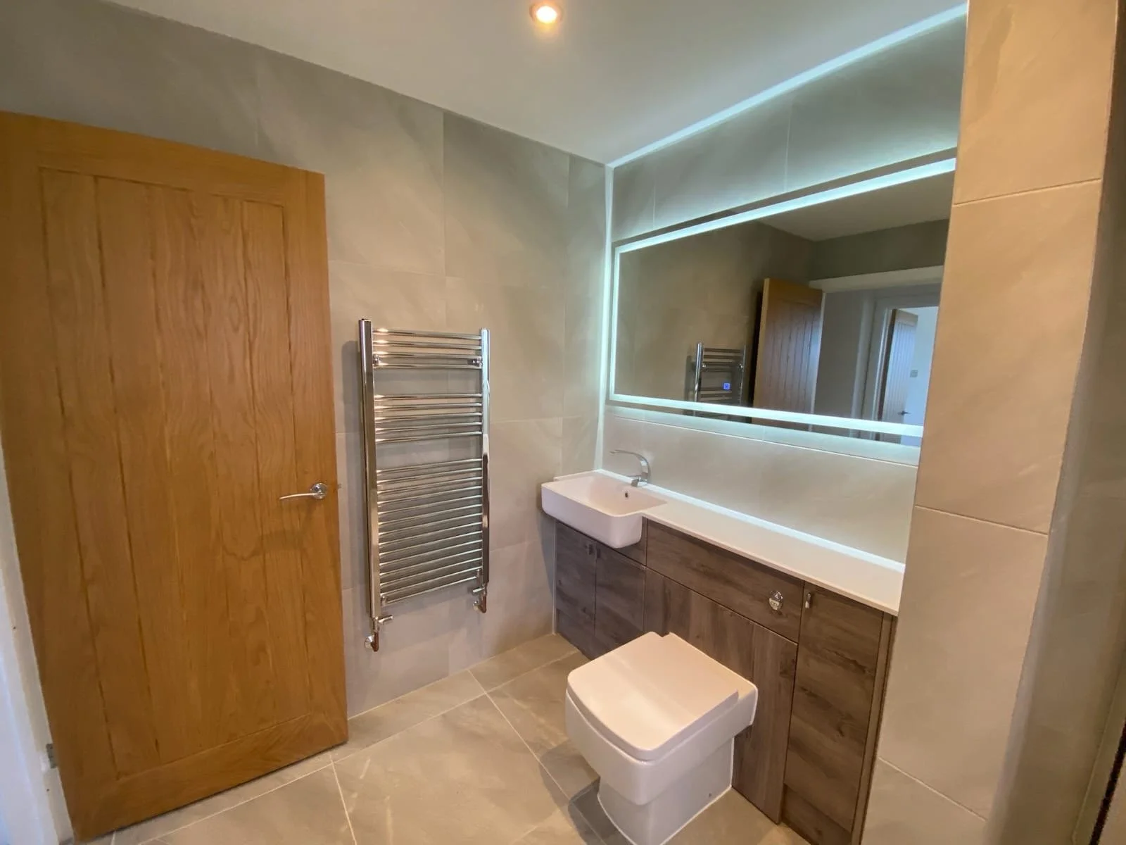 Modern bathroom with wooden door, wall-mounted towel rack, large mirror with backlit LED lighting, white sink, and wooden vanity cabinet.