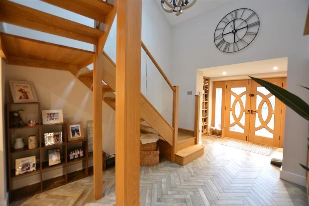 Entryway with wooden staircase, large wall clock, wooden front door with glass panels, and shelves with framed photographs and decorations.