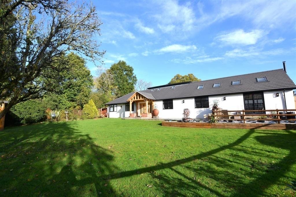 A large white house with a dark gray roof, multiple skylights, black window bars, and a wooden porch, surrounded by a green lawn and trees under a partly cloudy blue sky.