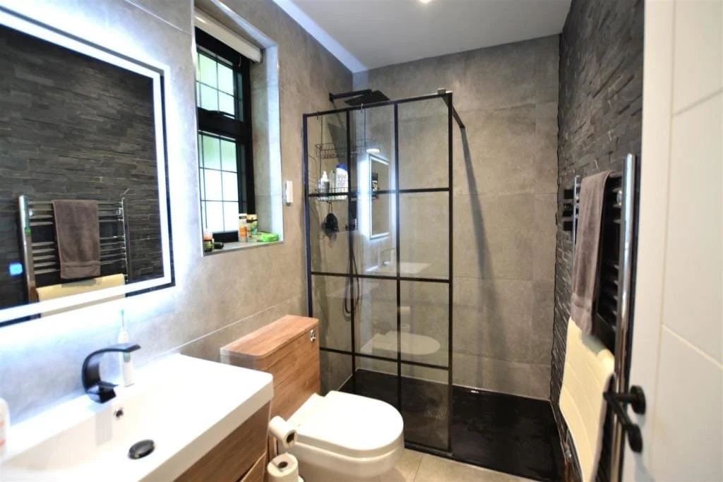 Modern bathroom with a walk-in shower, gray tiled walls, a white sink, and a small window.