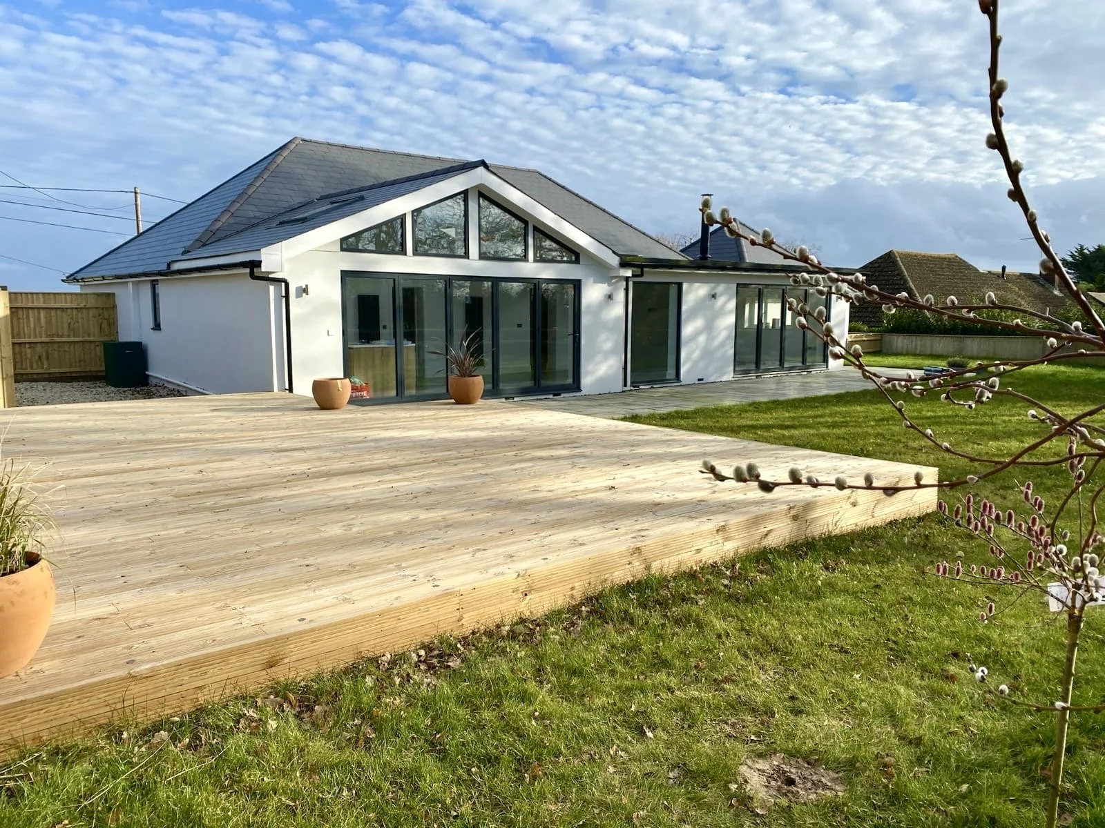 Modern white house with large sliding glass doors, a wooden deck, a green lawn, and a branch with budding flowers in the foreground.