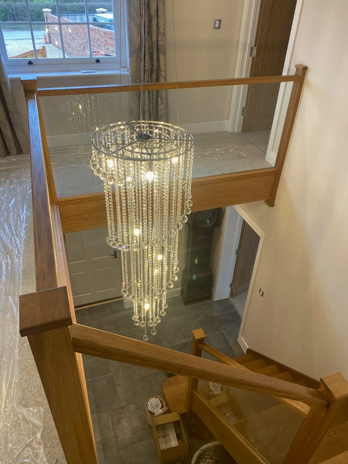 View of a chandelier hanging from the ceiling of a two-story foyer, with a staircase, a front door, and a loft area with a glass railing, overlooking the foyer.
