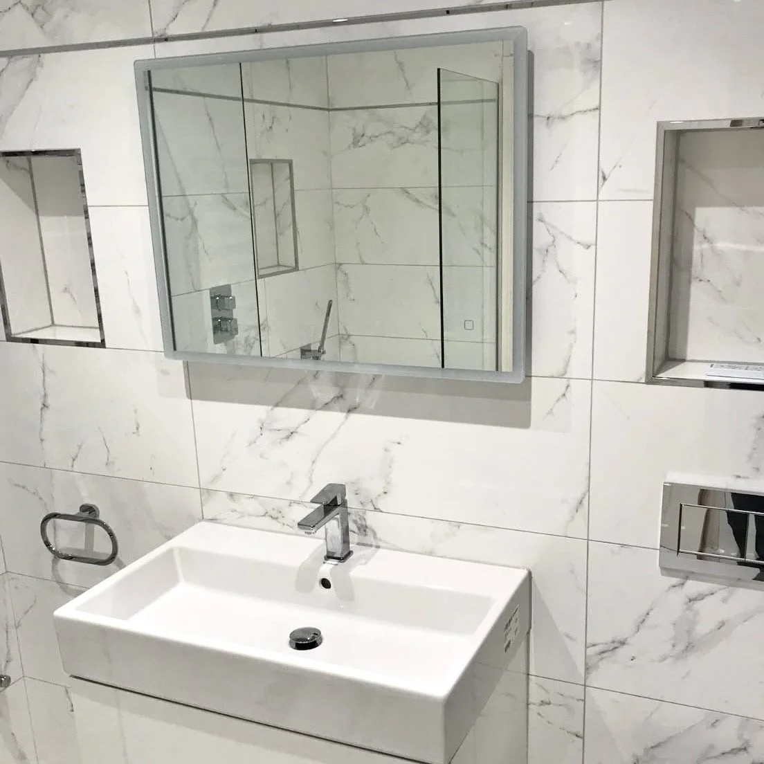 A modern bathroom features a white rectangular sink with a sleek chrome faucet, marble tiled walls, a mirror reflecting the shower area, and built-in wall niches for storage.