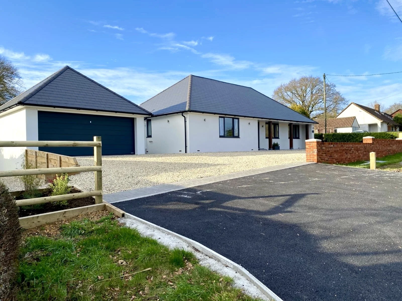 Modern house with white exterior walls, dark gray roof, garage, and driveway, situated in a suburban neighborhood with trees and neighboring homes in the background.