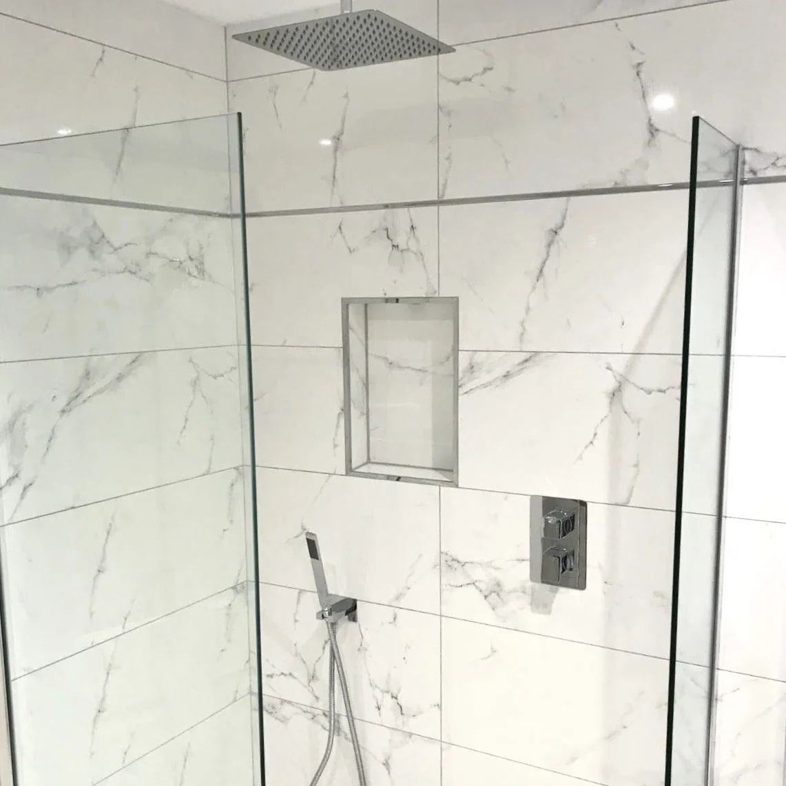 Shower with large white marble tiles, rainfall showerhead, built-in niche, and handheld shower handle.
