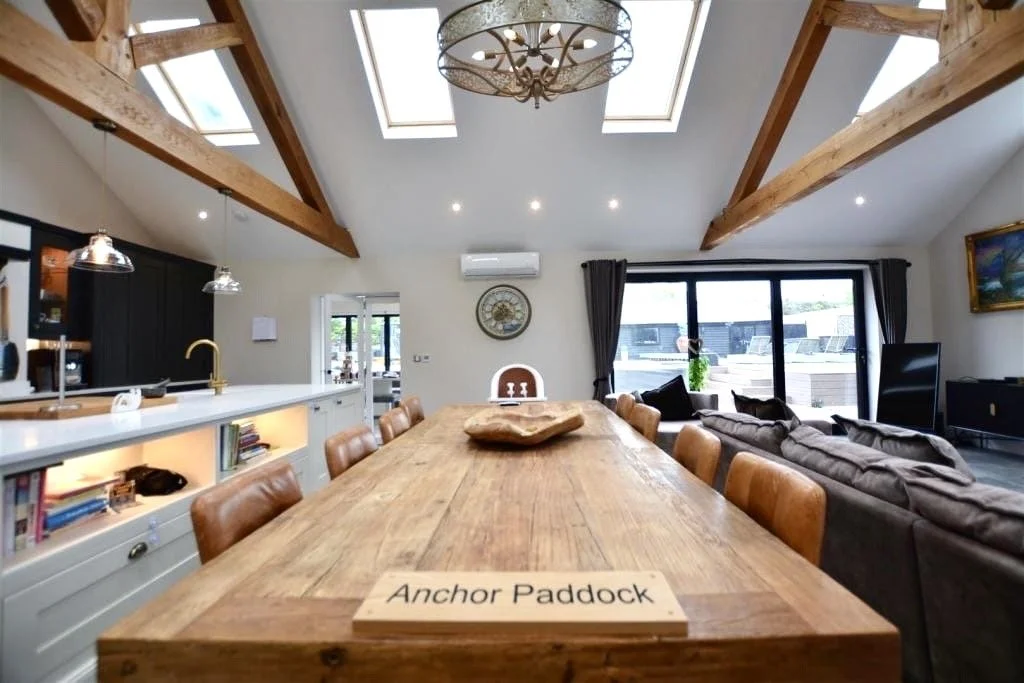 A spacious, open-concept living and dining room with a large wooden table labeled "Anchor Paddock," exposed wooden beams on the ceiling, skylights, and sliding glass doors leading to an outdoor patio.