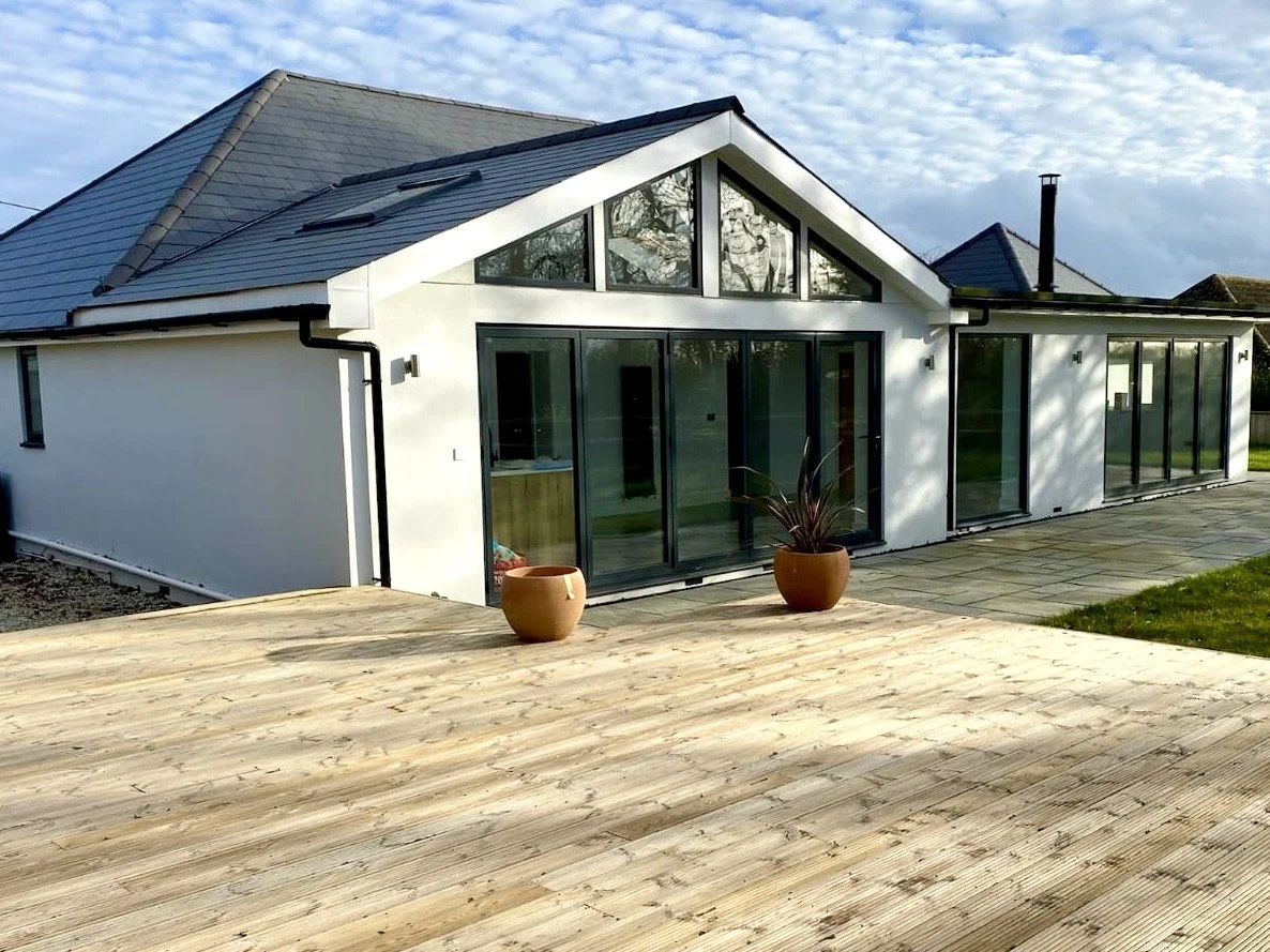 Modern house with large glass sliding doors, a wooden deck, and potted plants outside. The house has a white exterior and a triangular roof with skylights.