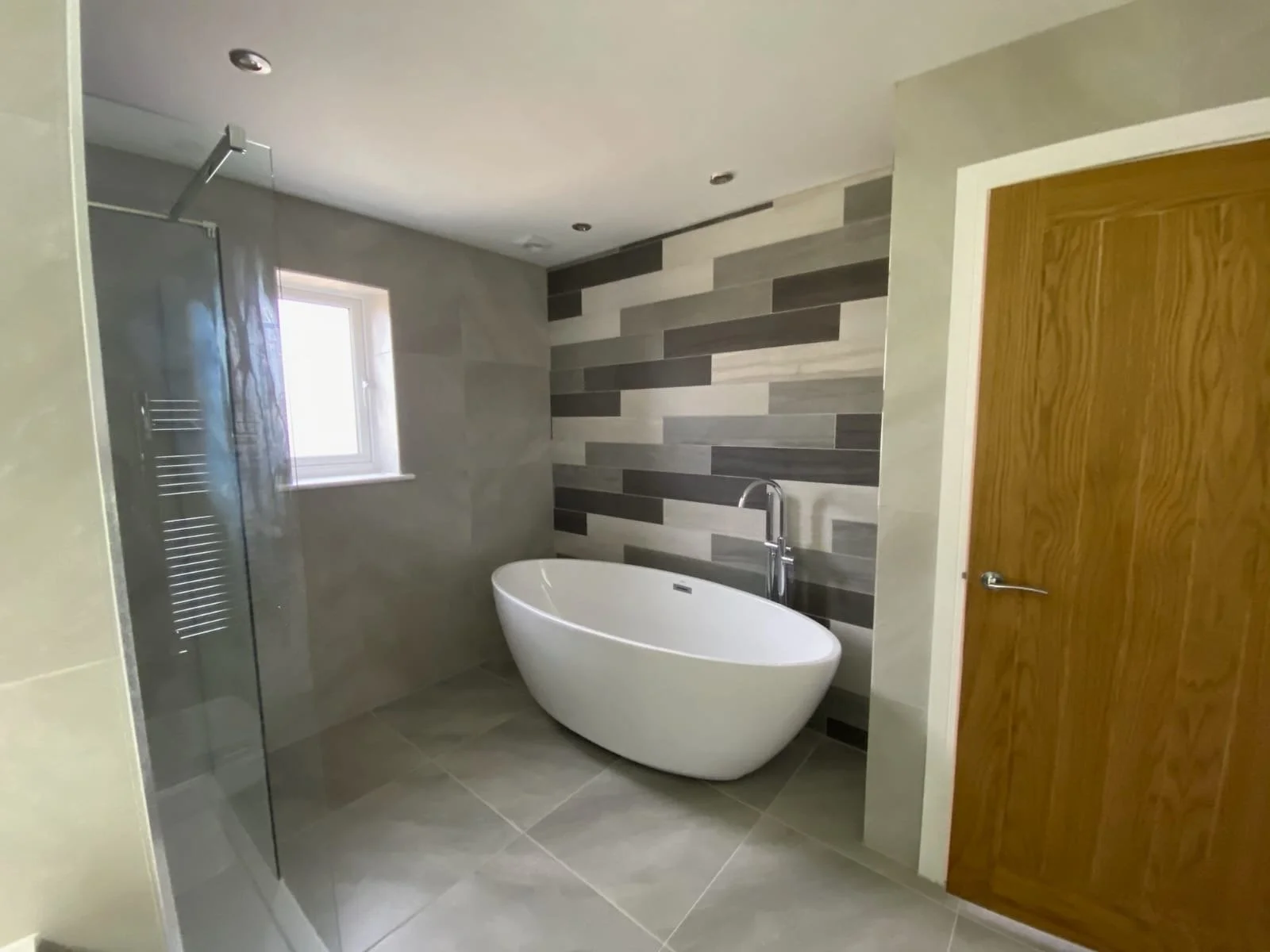 Modern bathroom with free-standing bathtub, wood door, glass shower enclosure, small window, and tiled walls in shades of gray.