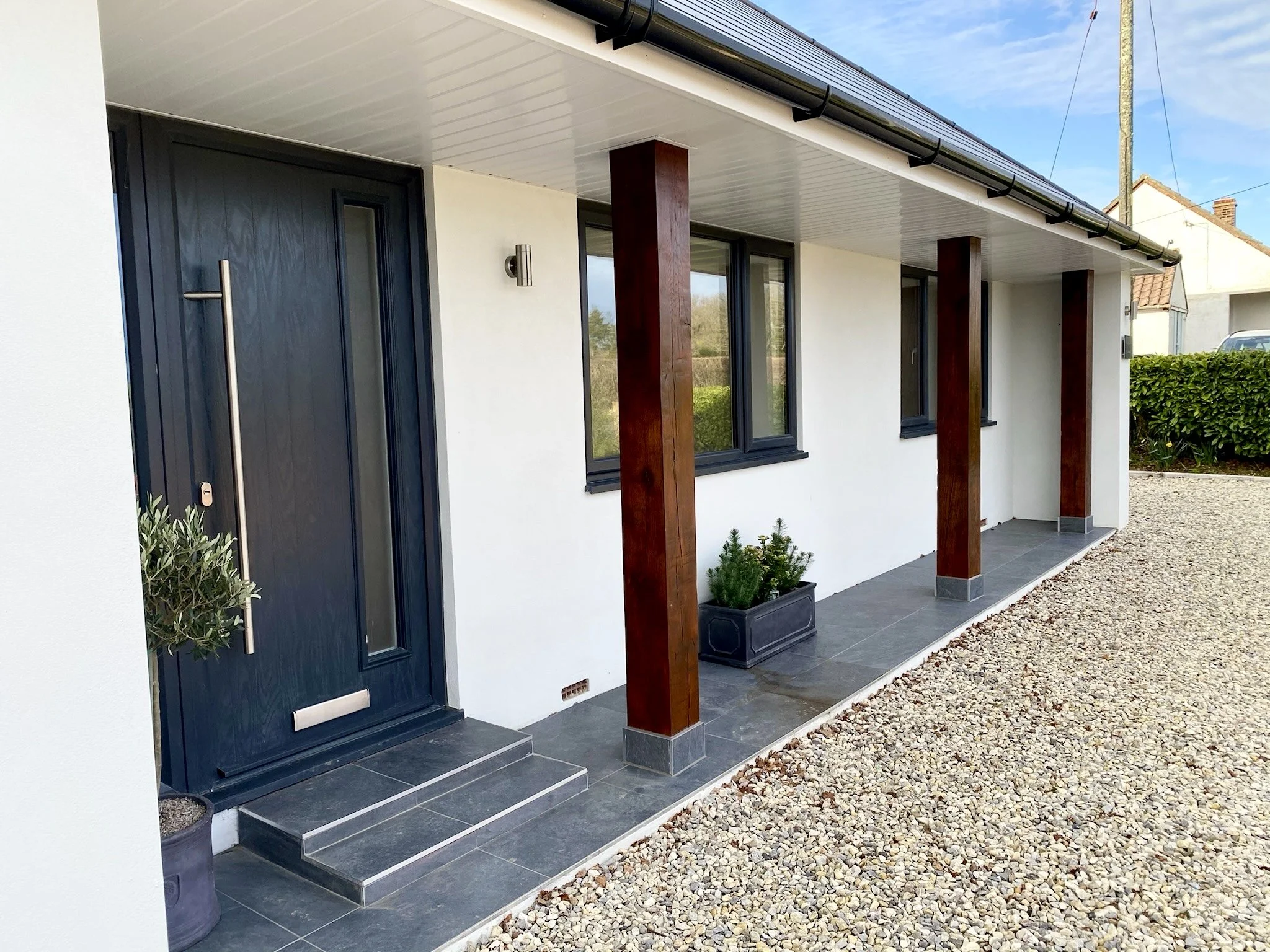 Front entrance of a modern house with dark blue door, wooden columns, grey tiled porch, small potted plants, and gravel driveway.