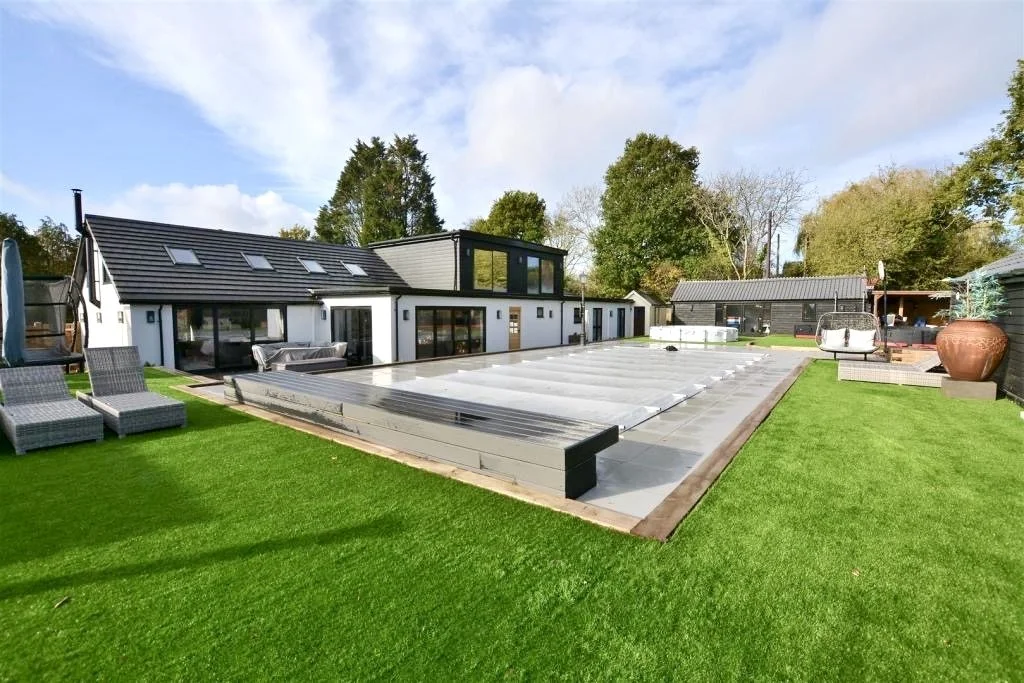 Modern backyard with a swimming pool covered by a protective cover, surrounded by well-maintained green grass, outdoor lounge chairs, and contemporary house with large windows and patio furniture, under a partly cloudy sky.