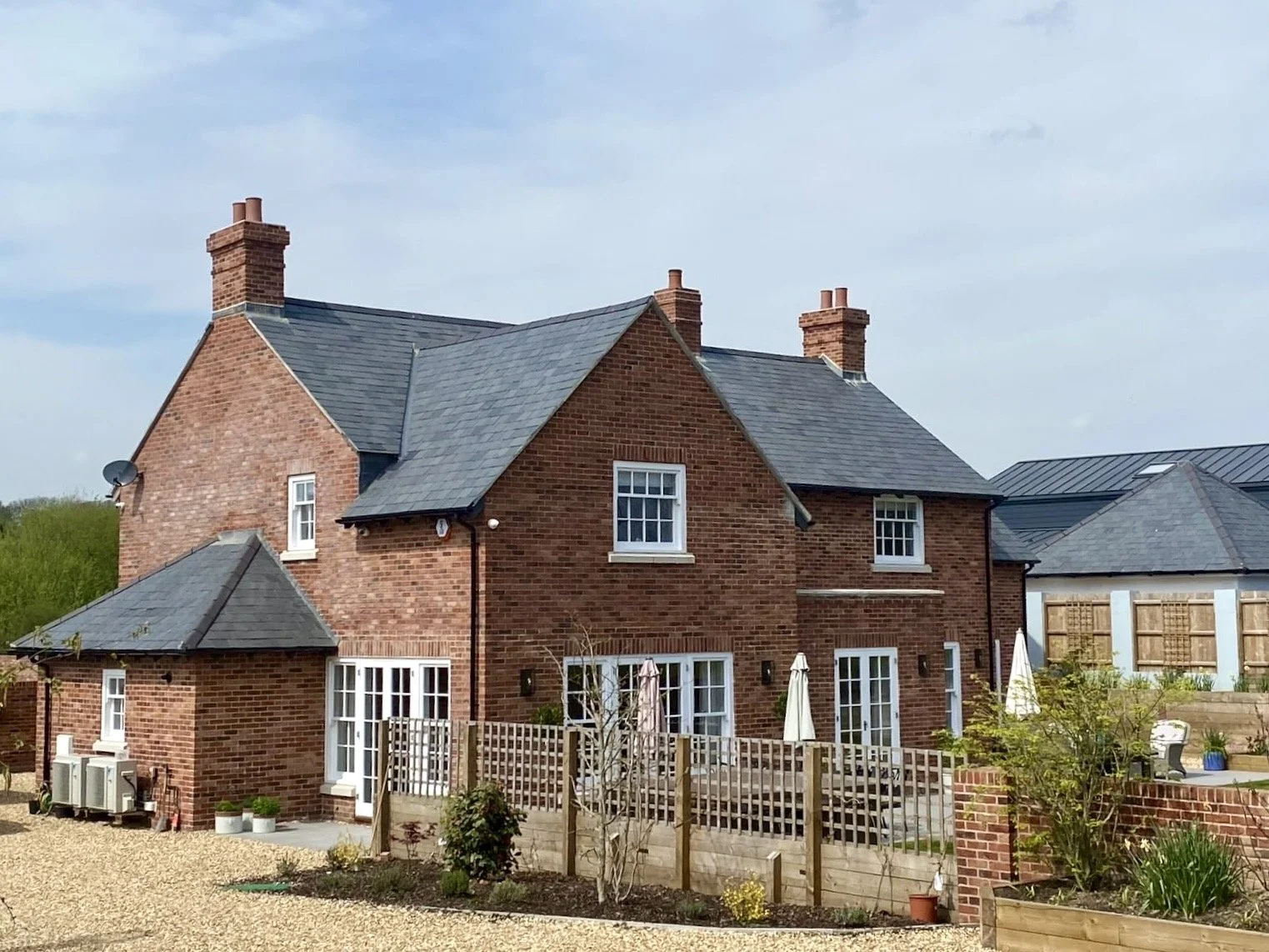 A red brick, two-story house with multiple chimneys, several windows, a small extension, and a garden with outdoor furniture and potted plants.