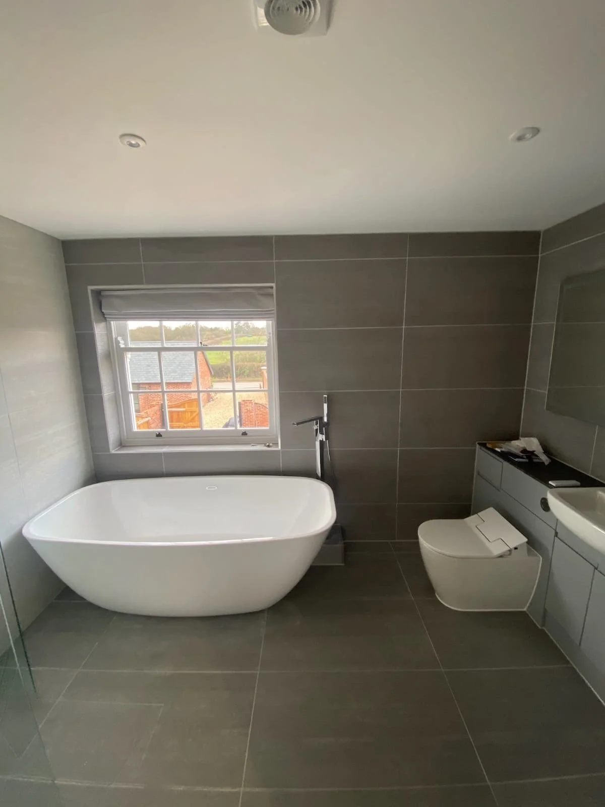 Modern bathroom with a freestanding bathtub, gray tiled walls, a window showing some houses outside, and a toilet with a closed lid.