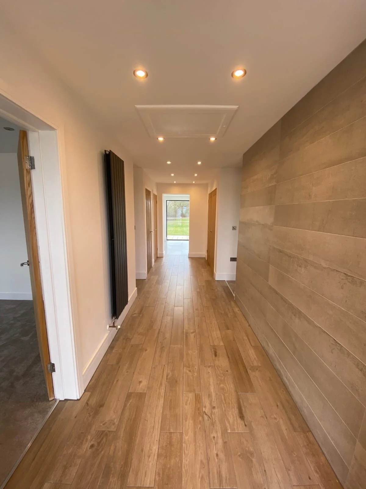 Empty hallway with wooden flooring, white walls, ceiling lights, and a large black radiator on the left, leading to a door or window at the end showing a green outdoor scene.