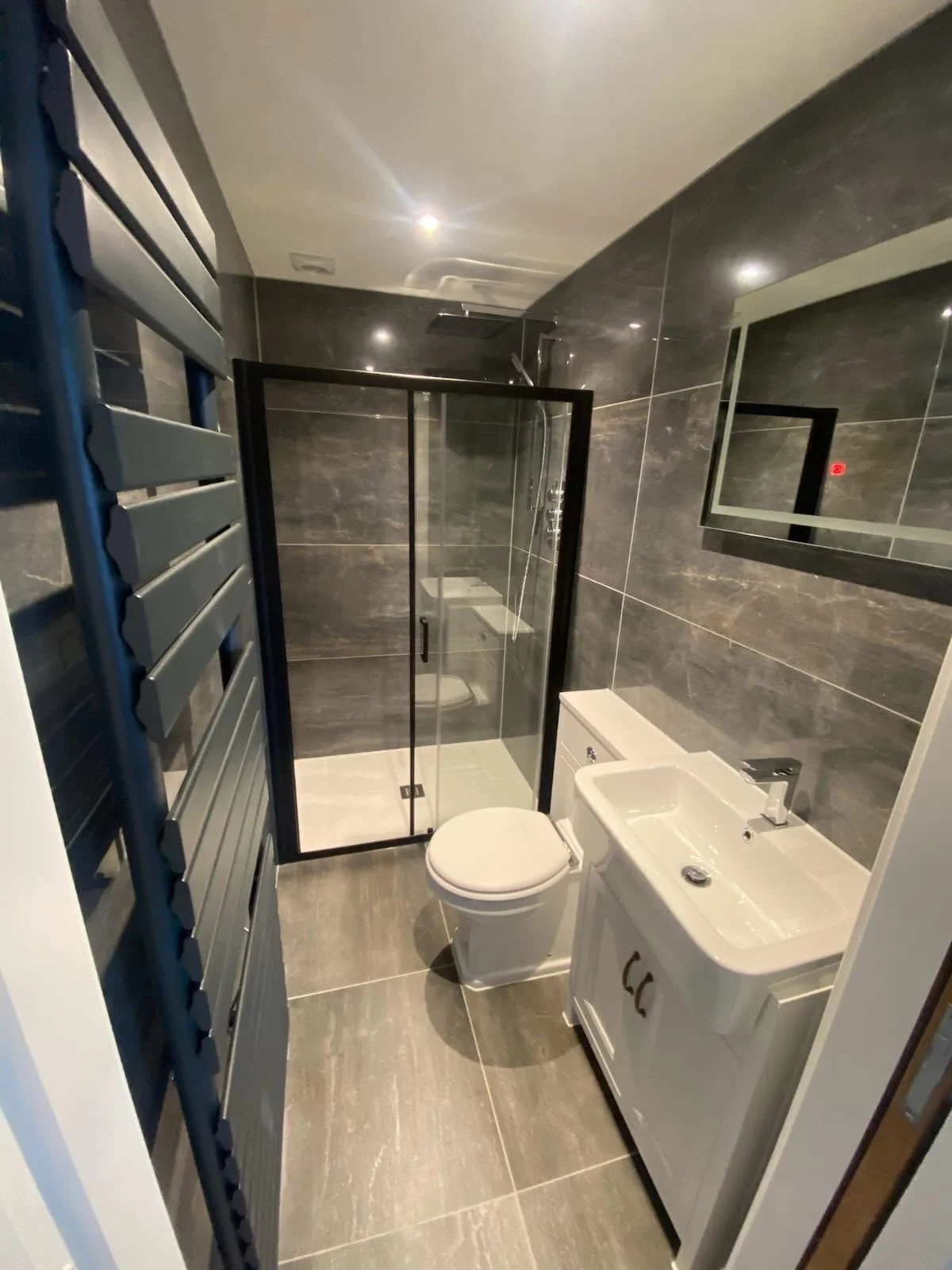 Modern small bathroom with gray tiled walls and floor, glass shower enclosure, white toilet, and white vanity with sink.