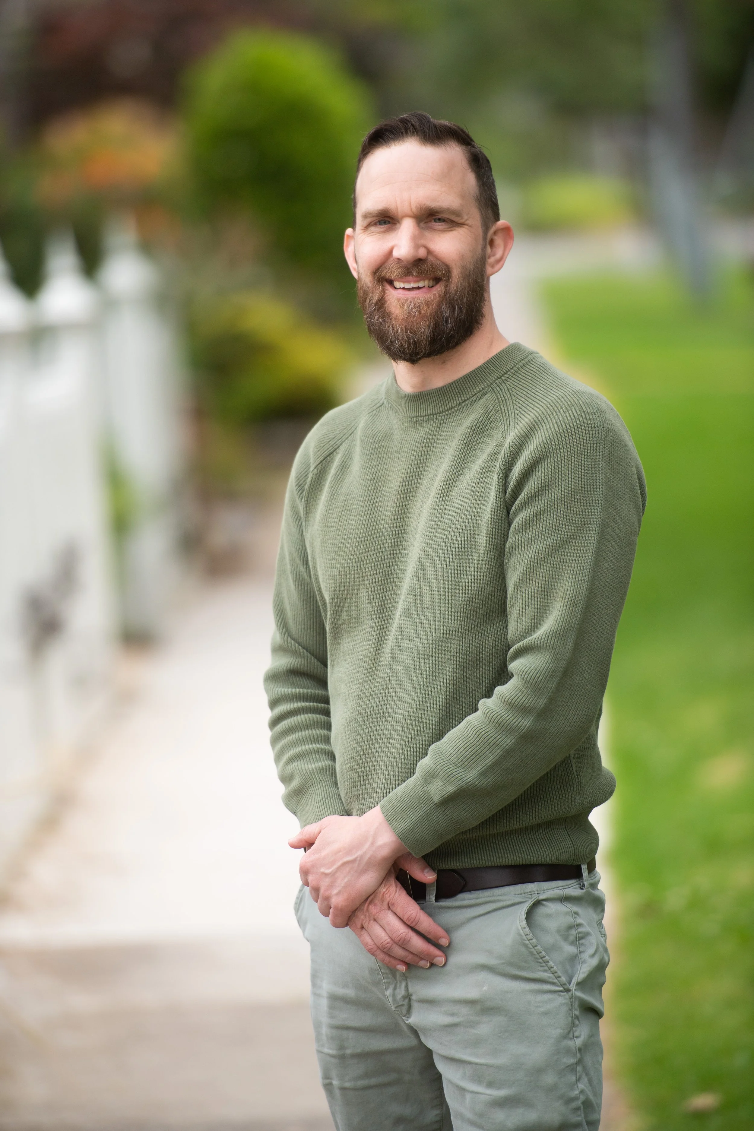 An image of Dr. Brendan McLaren, Clinical neuropsychologist, smiling outdoors, wearing a green sweater and light gray pants, standing on a sidewalk with a white fence and green grass in the background.
