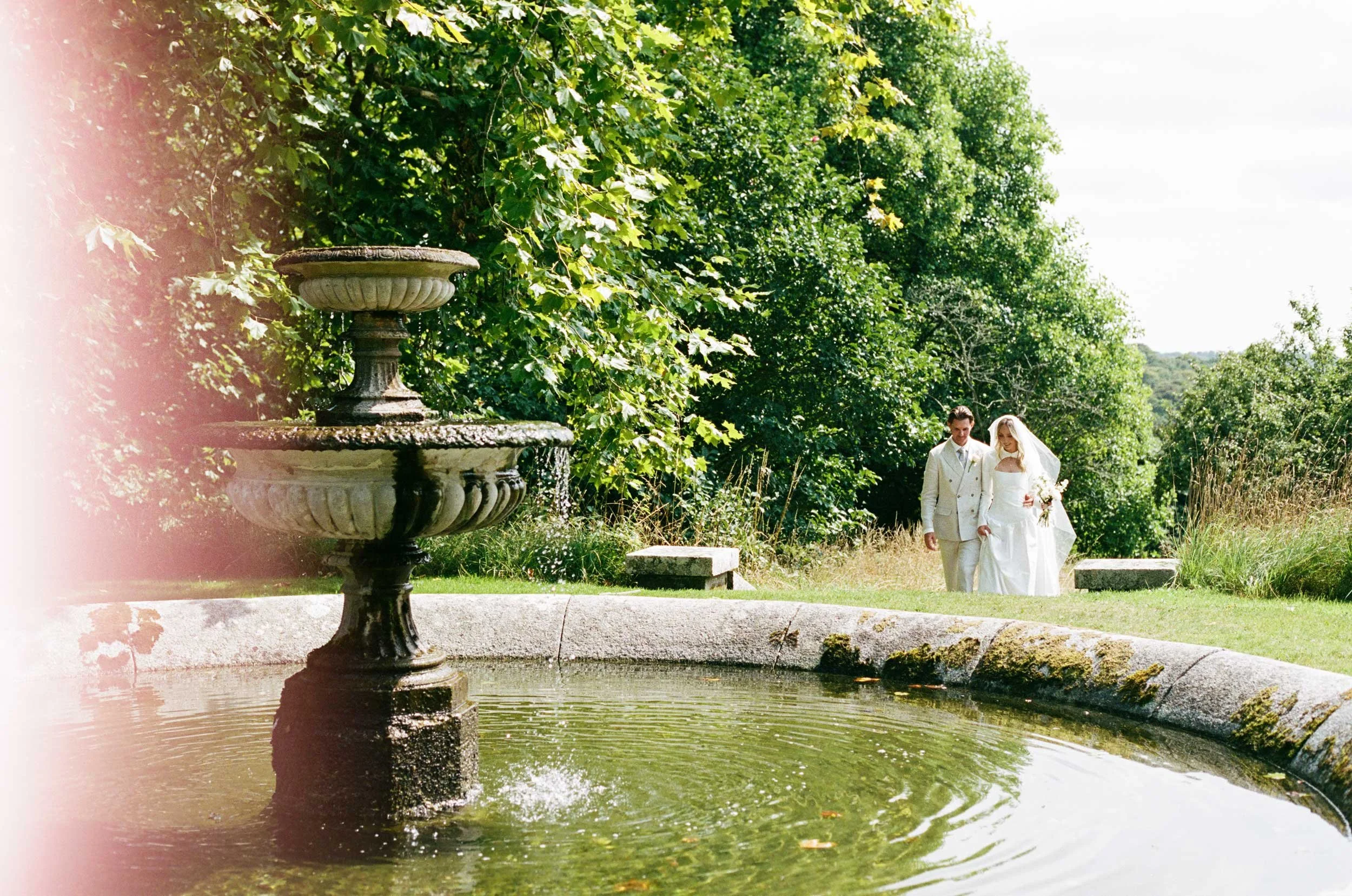 35mm film wedding photography of couple walking beside garden fountain, natural documentary moment in Cornwall