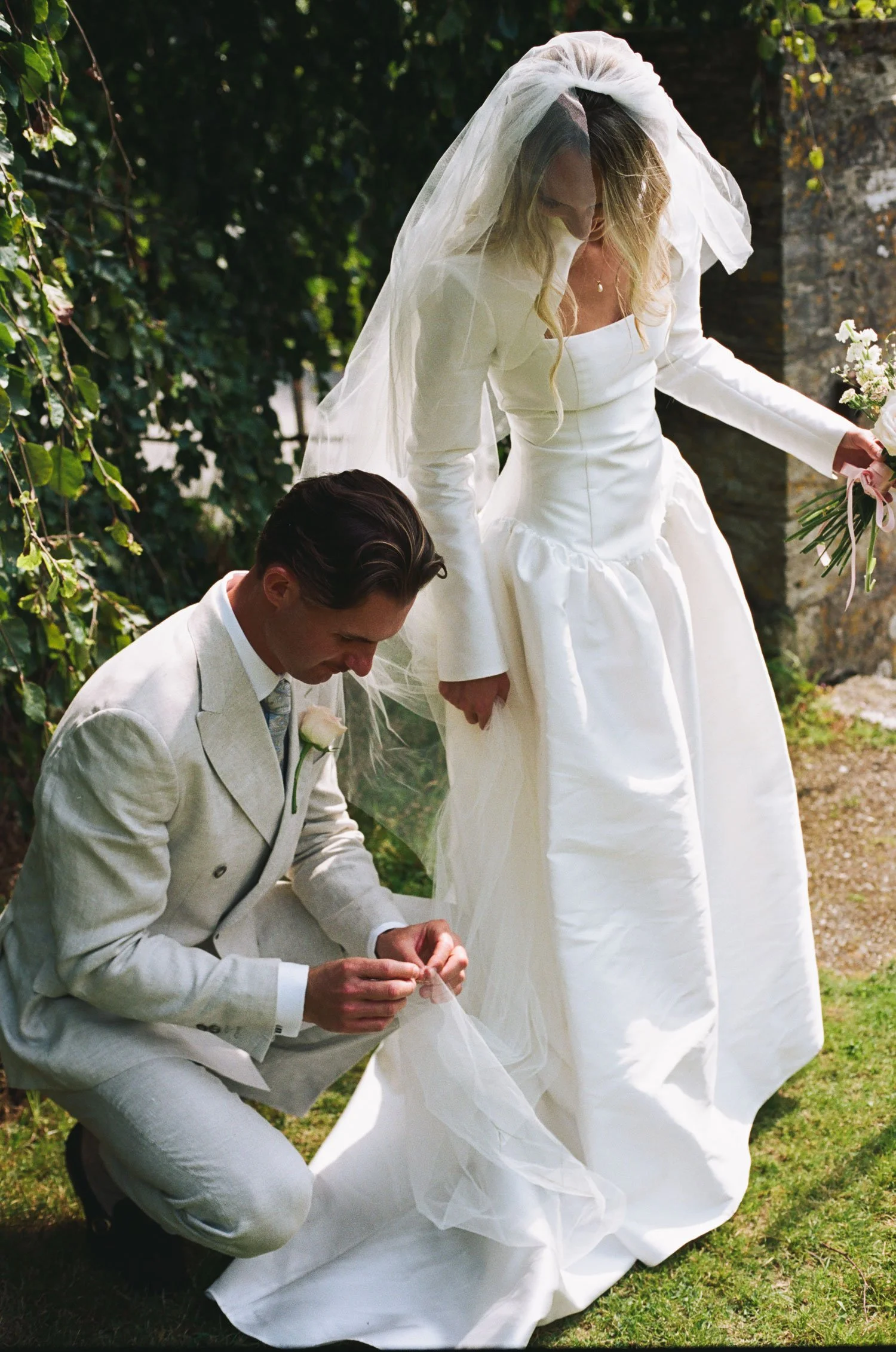 35mm film wedding photograph of groom adjusting bride’s dress outdoors, natural documentary moment