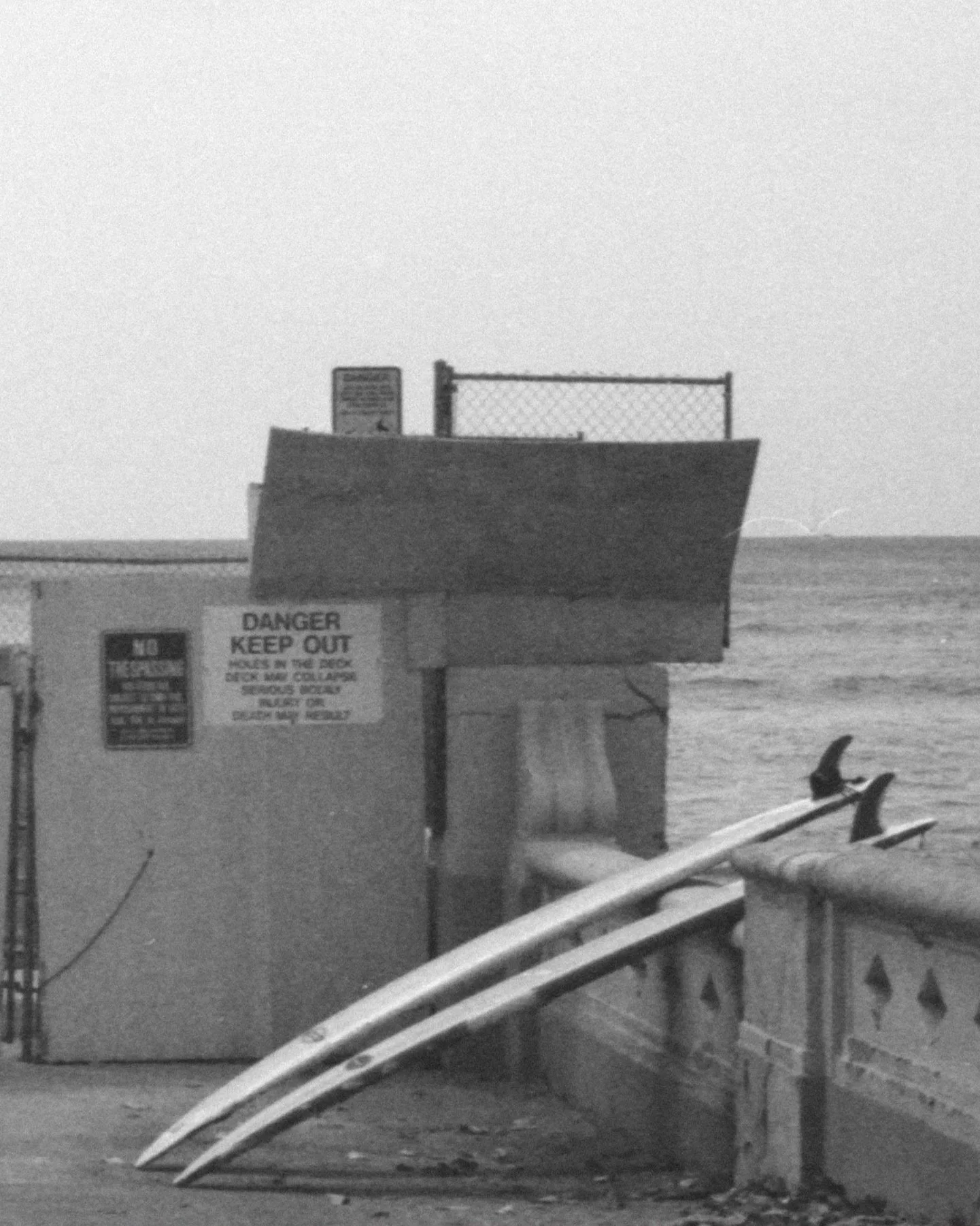 Black and white film photograph of surfboards leaning against seafront wall, editorial analogue work by Karl Mackie