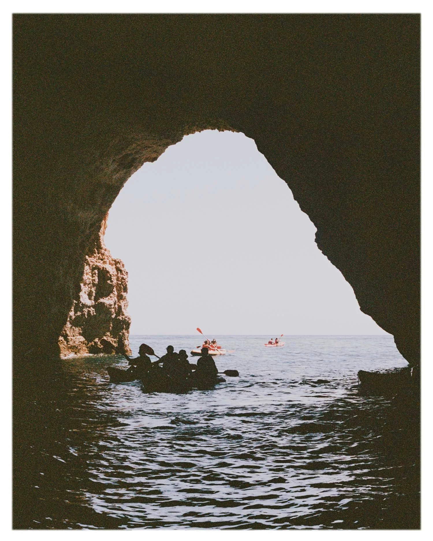 Colour film photograph of kayakers silhouetted inside sea cave opening, analogue travel and lifestyle imagery by Karl Mackie