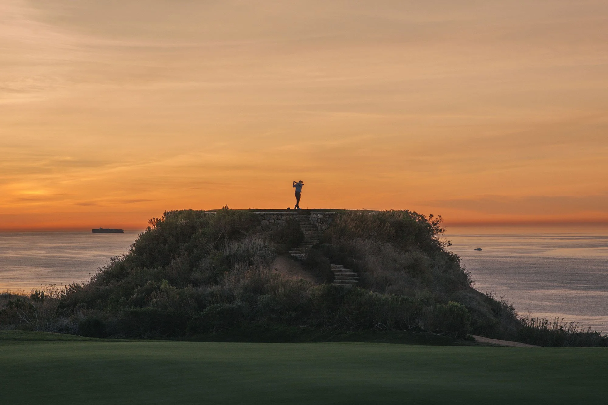 Golfer silhouetted against an orange sunset on a coastal golf course overlooking the Pacific Ocean