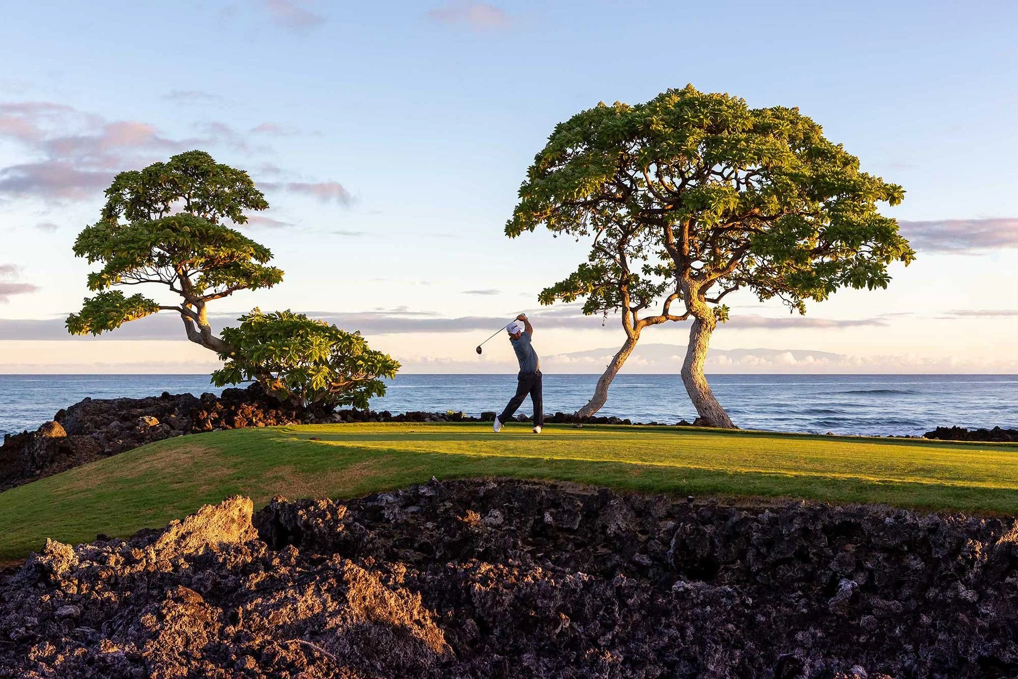Golfer walking beneath trees beside the ocean at Four Seasons Golf Resort Hawaii