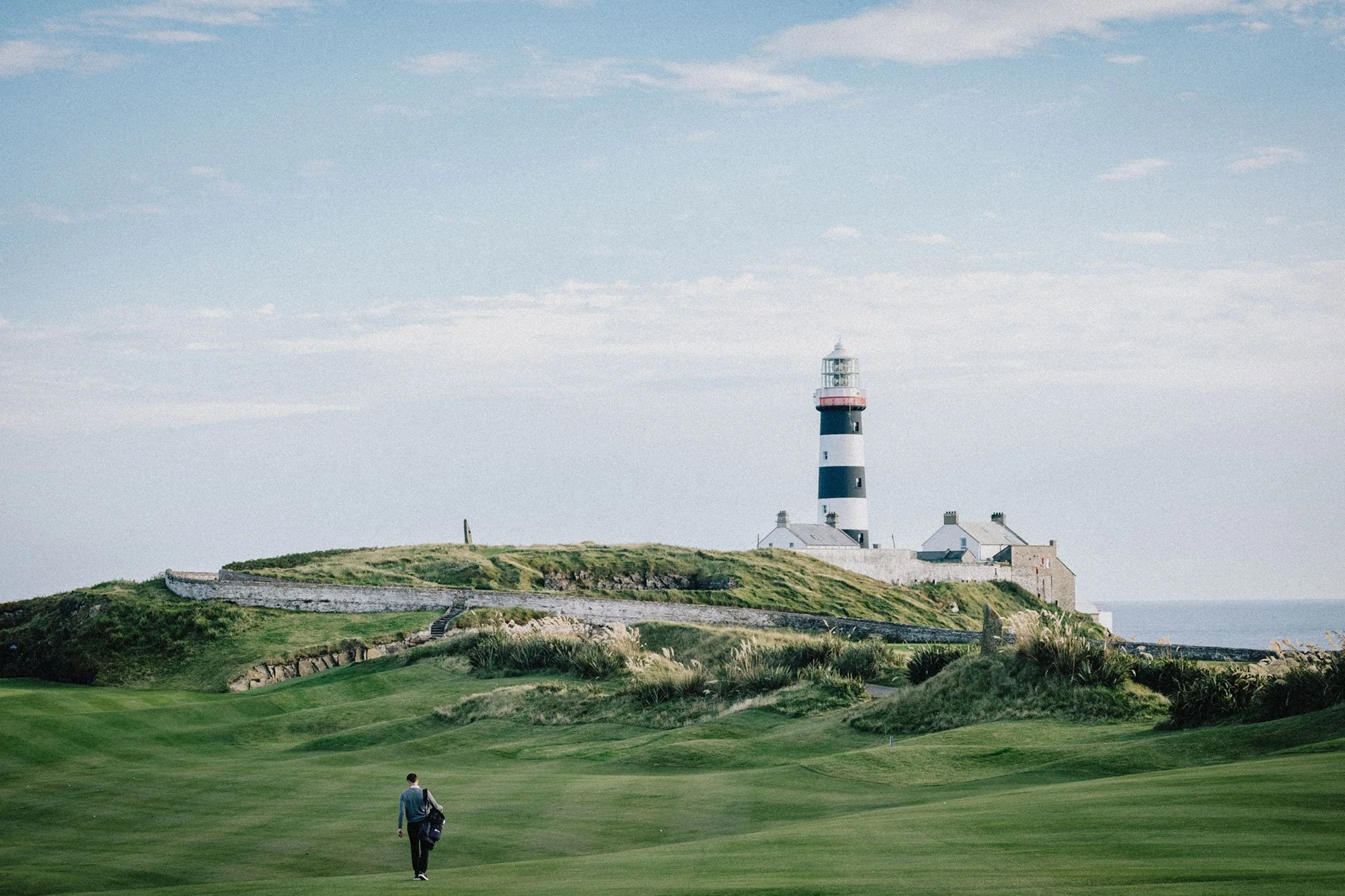 Golfer walking across the fairway at Old Head Golf Links in Ireland beneath the iconic black-and-white lighthouse