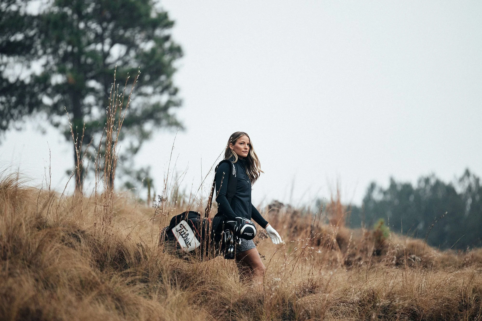 Female golfer carrying clubs through wild grass on a woodland course