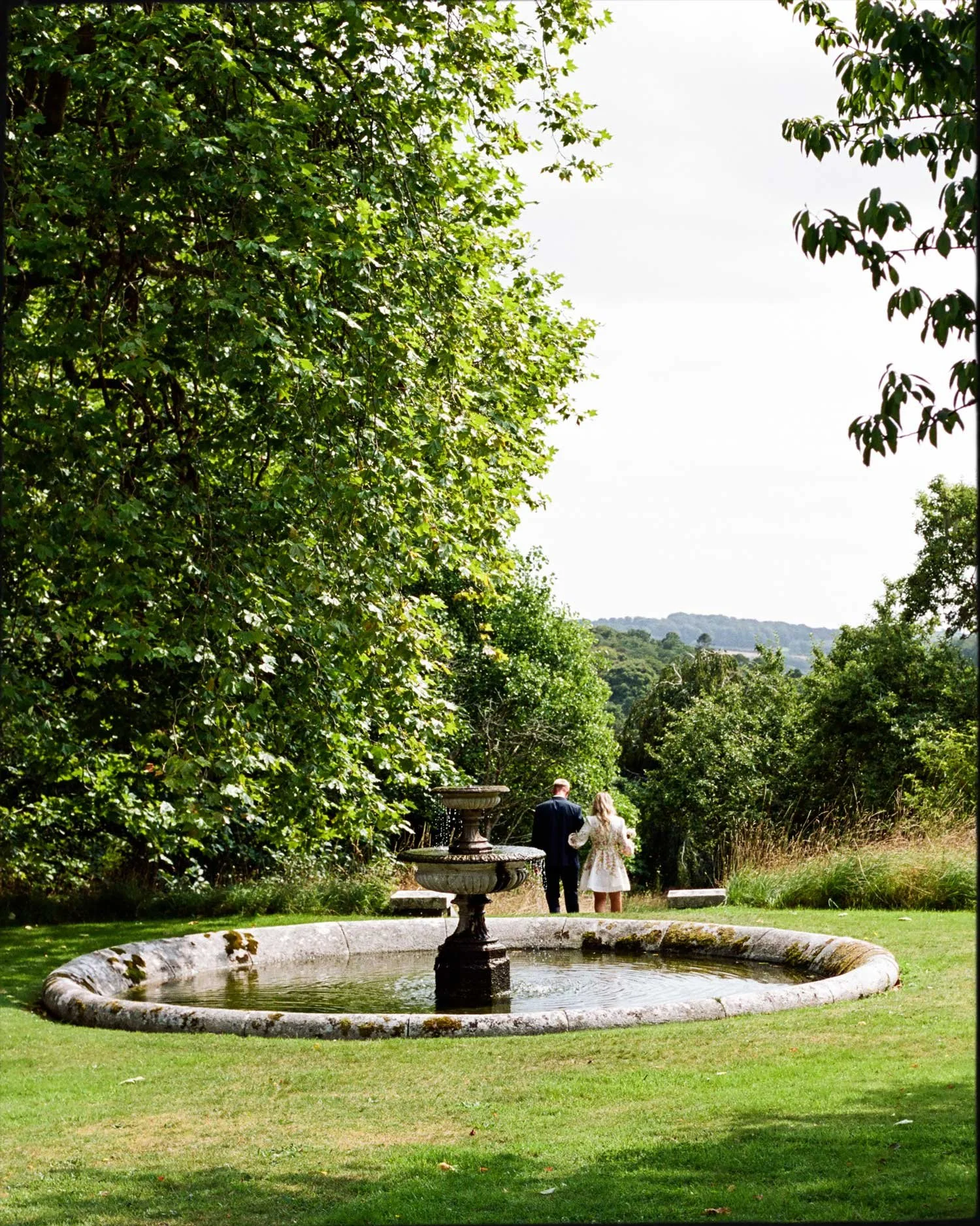 Documentary wedding photography on 35mm film, couple walking beyond garden fountain in Cornwall