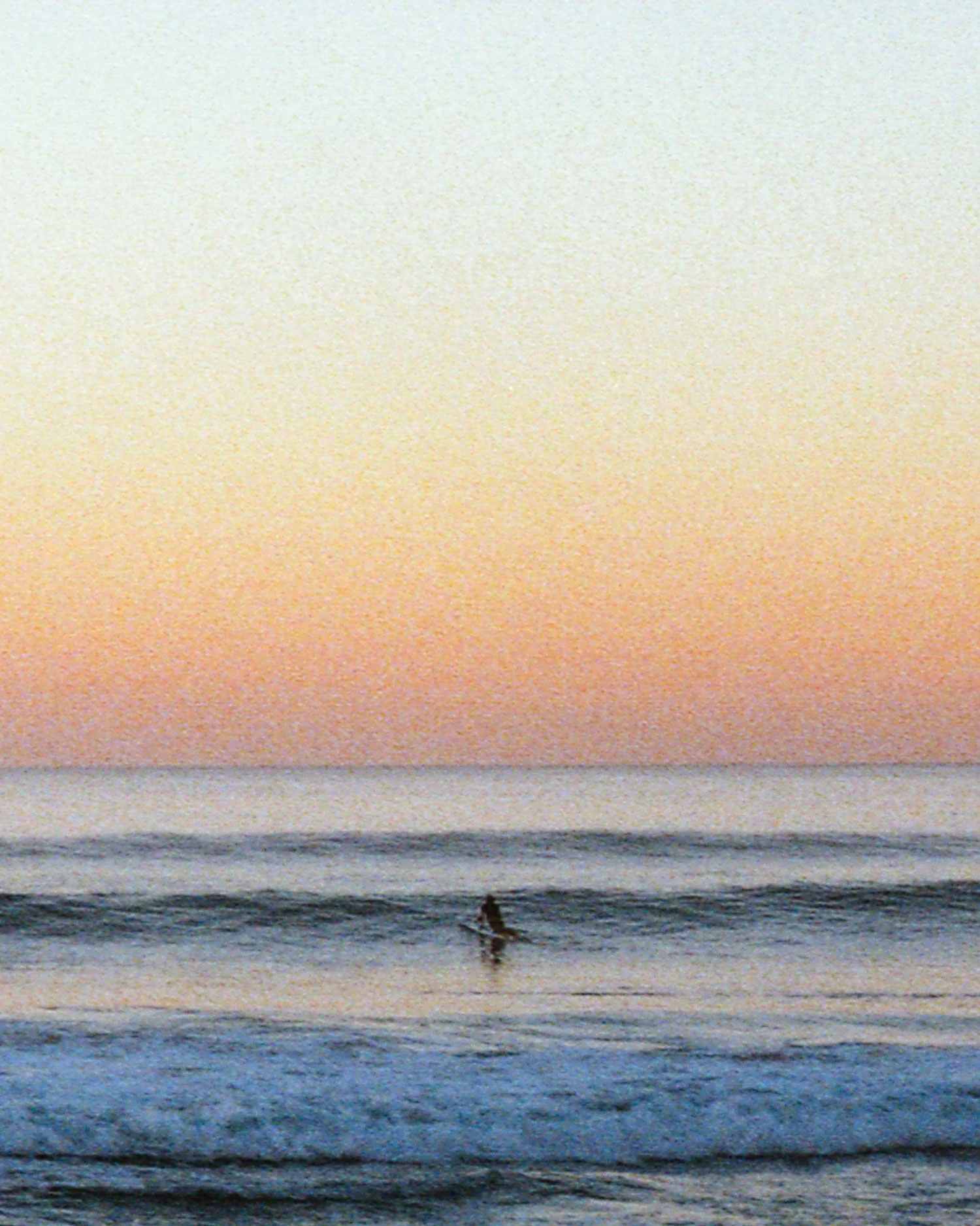 Soft pastel sunset over Watergate Bay in Cornwall with a lone surfer waiting beyond the shoreline. Shot on film by Karl Mackie.