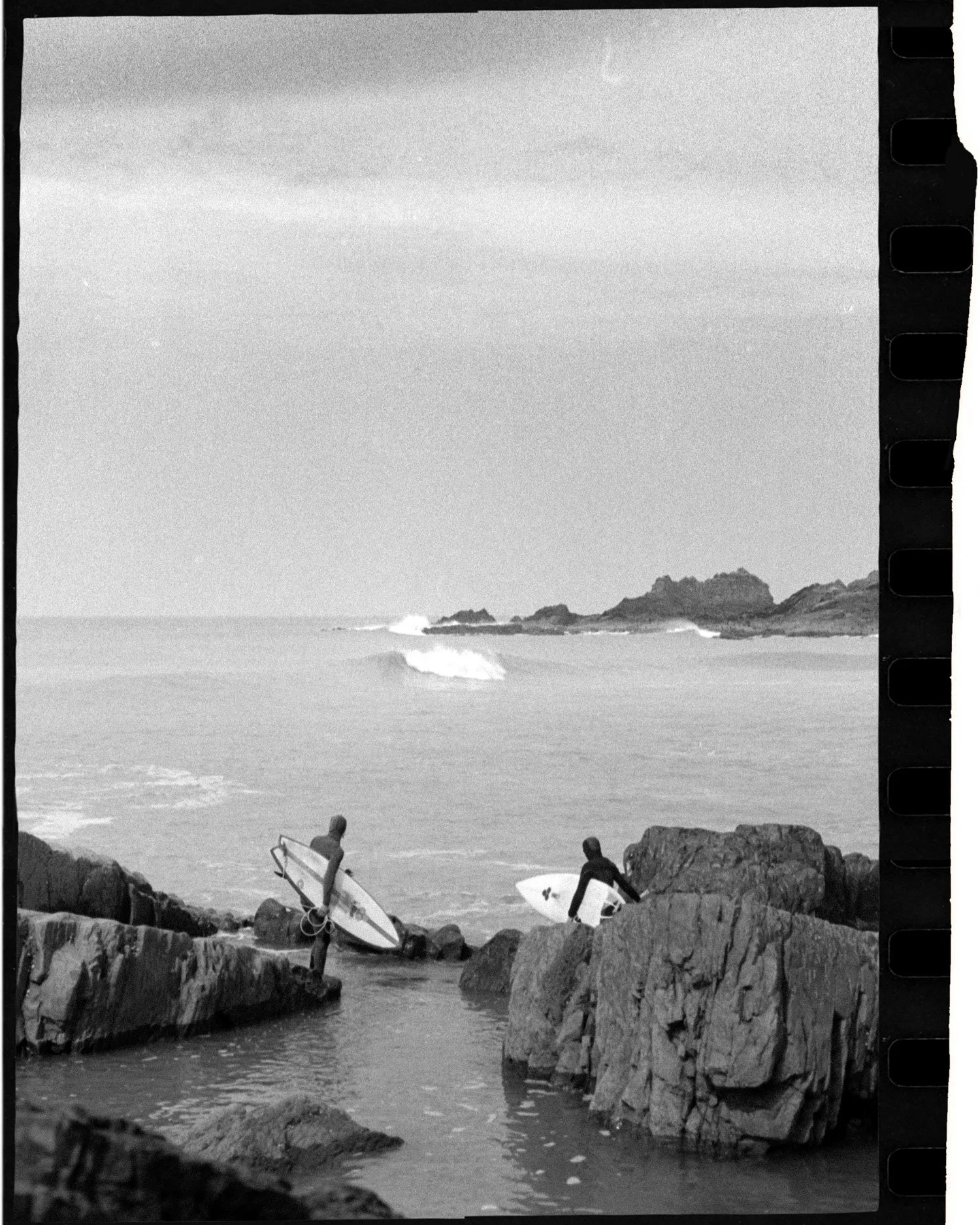 Black and white 35mm film photograph of surfers standing on rocky coastline, analogue editorial work by Karl Mackie