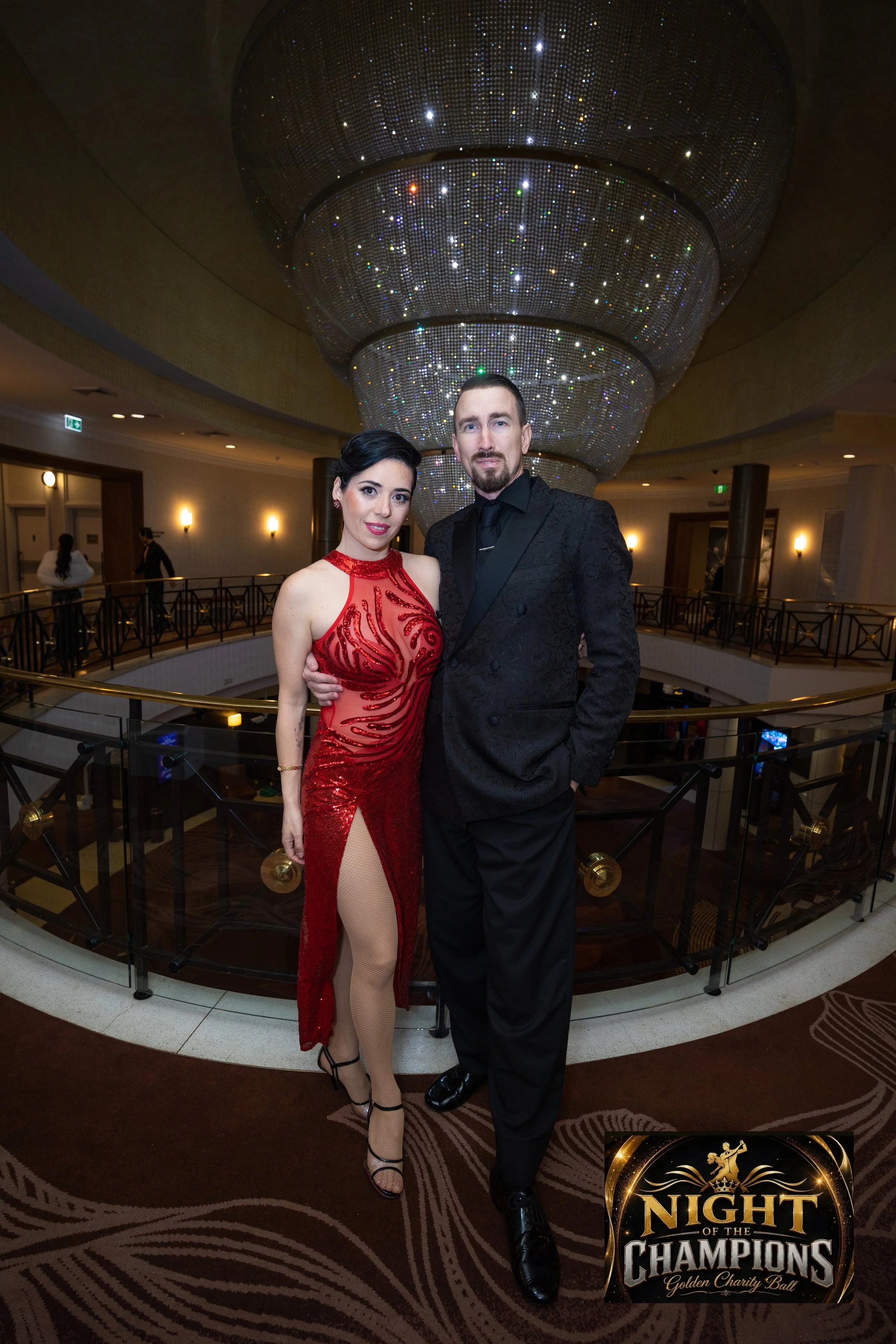 A man and a woman dressed in formal attire, standing together on a balcony inside a venue with a large, ornate chandelier overhead. The woman is wearing a red, sequined gown with a slit and beige fishnet stockings, while the man is in a black suit. In the background, there are people walking and the logo for 'Night of the Champions' charity ball is in the corner.