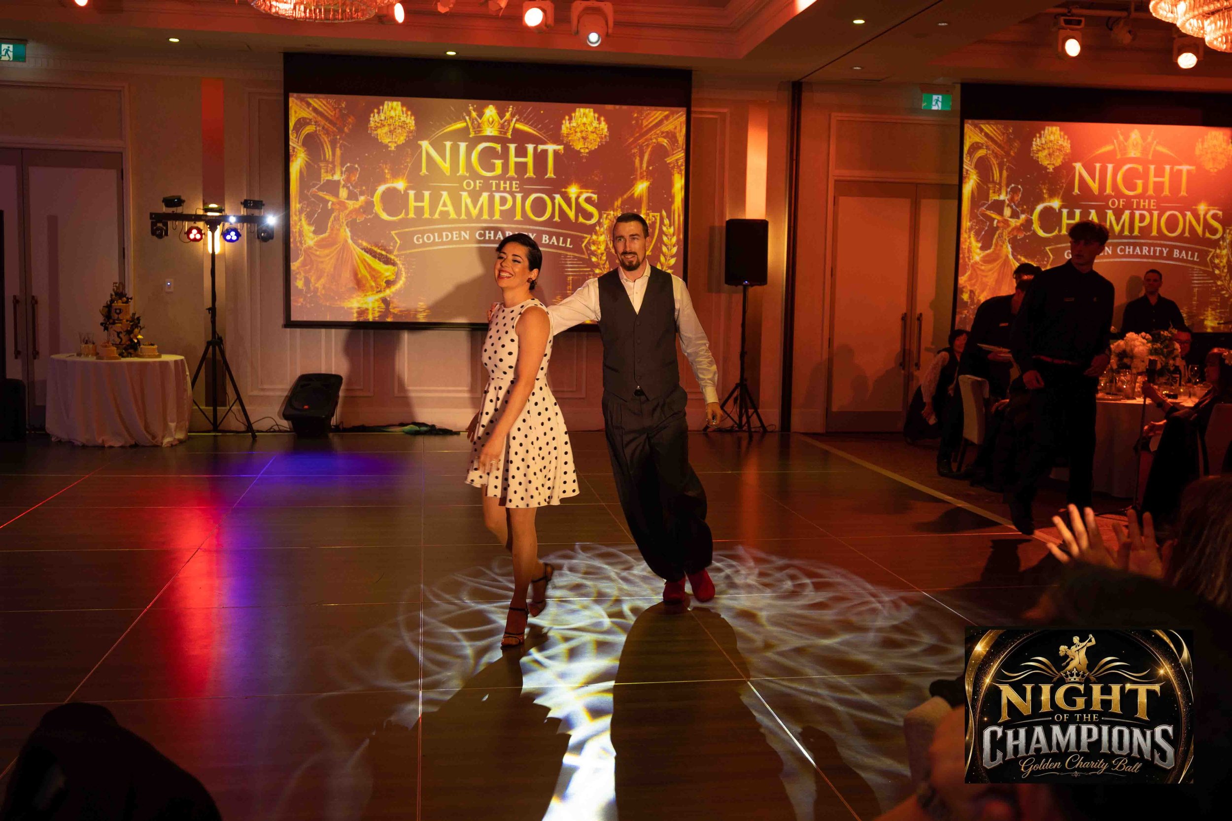 Two dancers, a woman in a polka dot dress and a man in a vest and dress shirt, are performing on a dance floor at a charity event called Night of the Champions. There is a large screen behind them displaying the event's name and logo, with chandeliers and guests seated at tables visible in the background.
