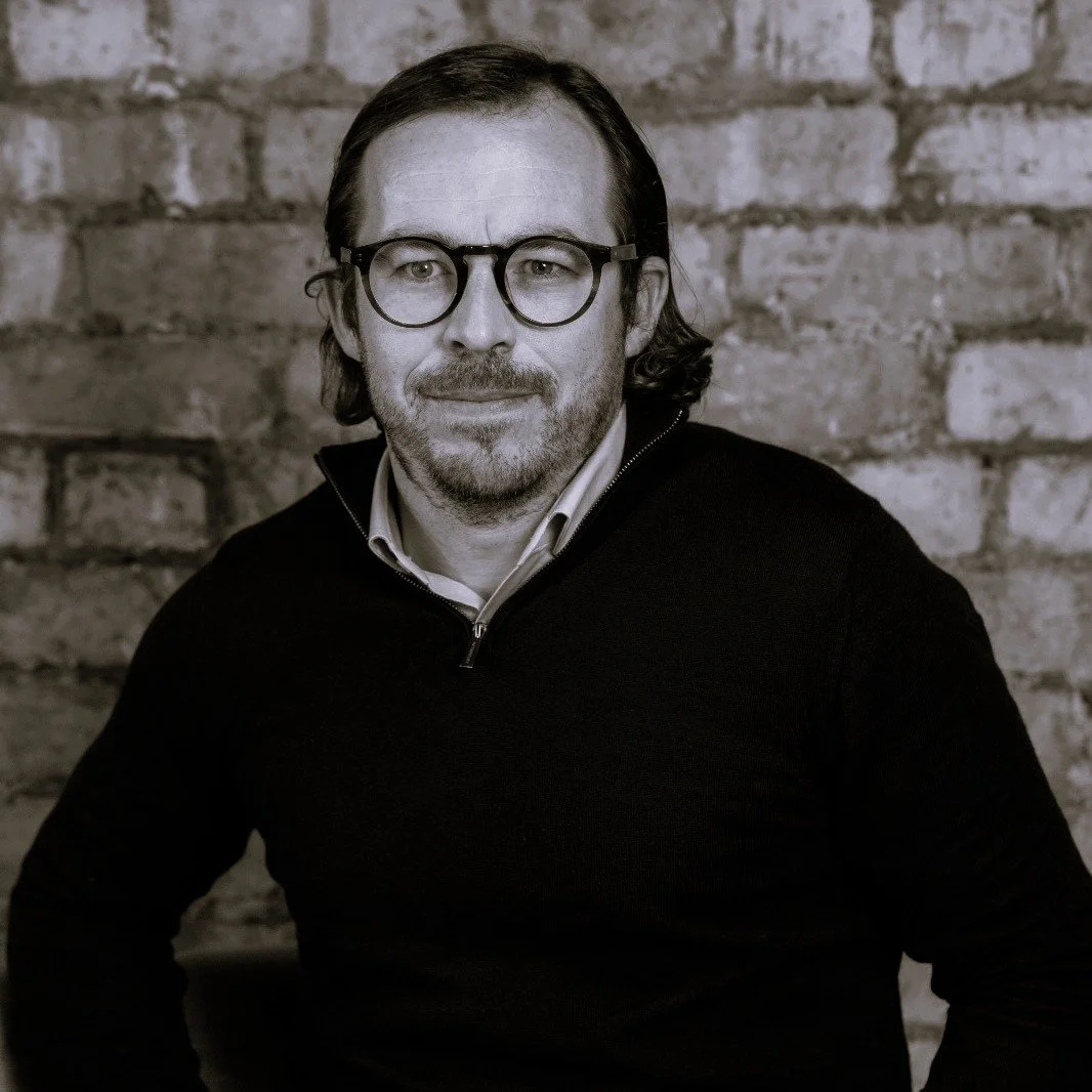 A man with medium-length hair, glasses, a beard, and mustache wearing a dark sweater and collared shirt, sitting against a brick wall.