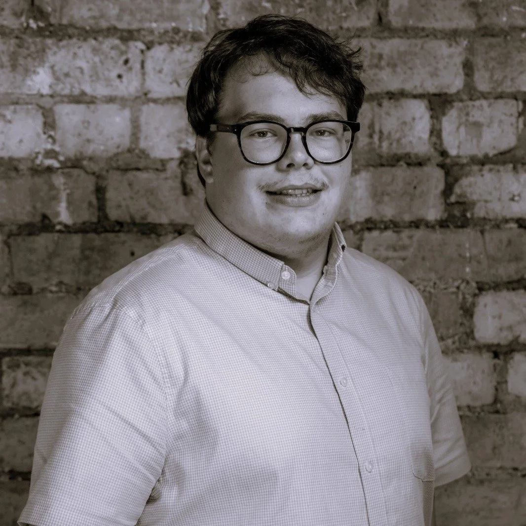 A young man with glasses and short hair, wearing a button-up shirt, standing against a brick wall.